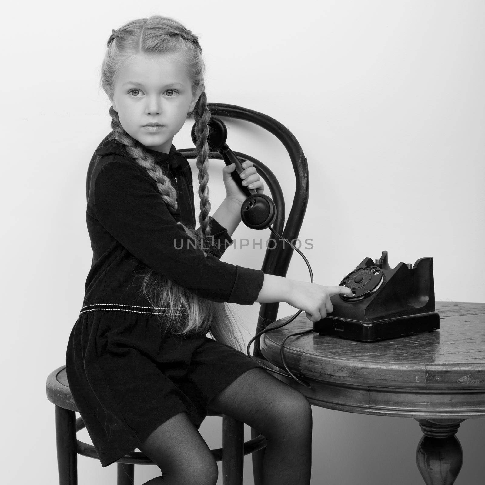 Smiling girl talking by old phone. Black and white shot of lovely kid sitting on chair in vintage room interior. Cute six years old kid speaking on vintage telephone