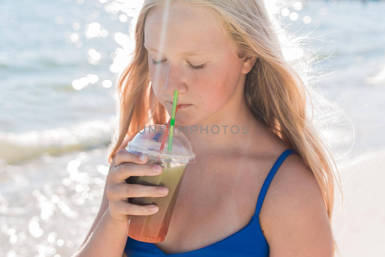 A young girl with blonde hair of European appearance, a teenager holds and drink a colored cold non-alcoholic cocktail in her hand against the background of the sea beach.