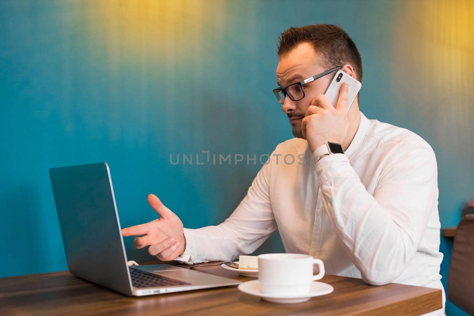 Portrait of a young attractive businessman in a white shirt, glasses, talking on the phone and looking at a laptop with a cup of coffee in a cafe.