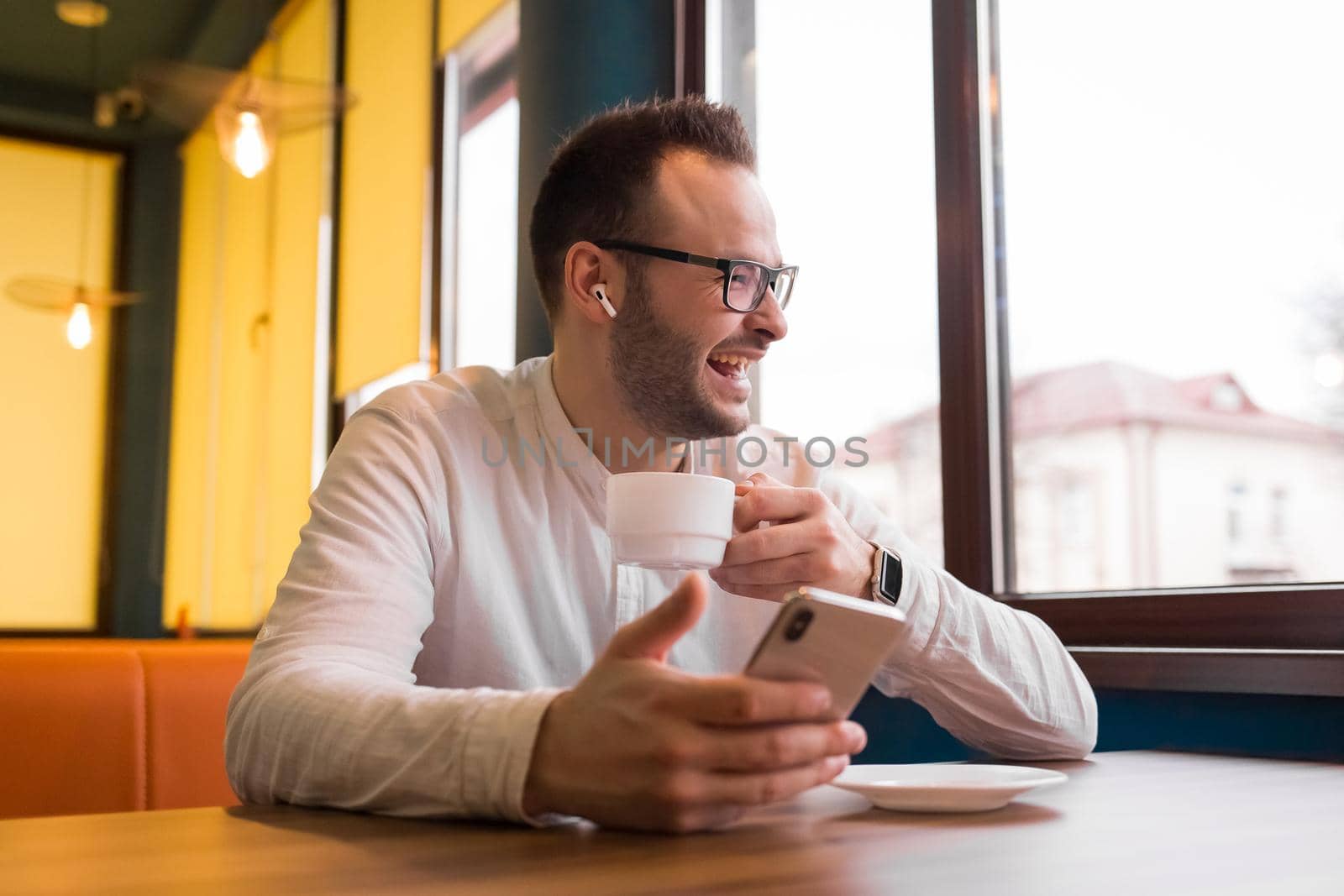 Portrait of a young handsome businessman in a white shirt, glasses, with a beard and headphones, spending time on the phone, drinking coffee and laughing.