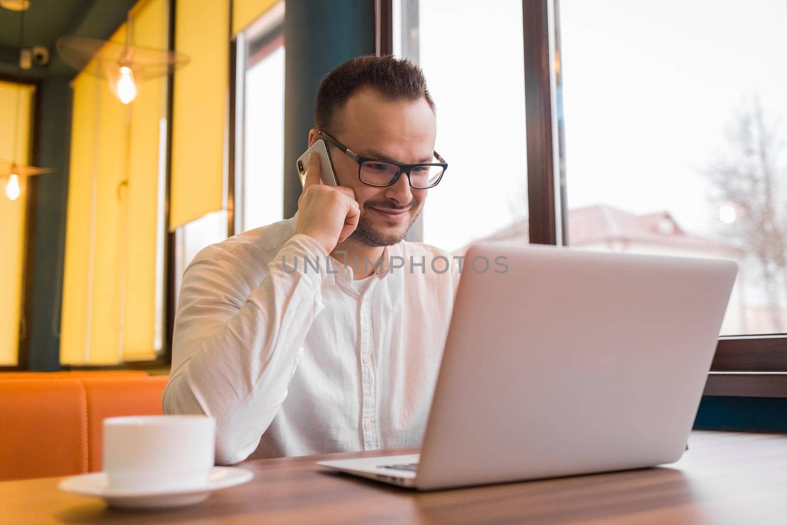 Portrait of a satisfied businessman in a white shirt, glasses, with a beard, talking on the phone and looking at a laptop with a cup of coffee in a cafe.