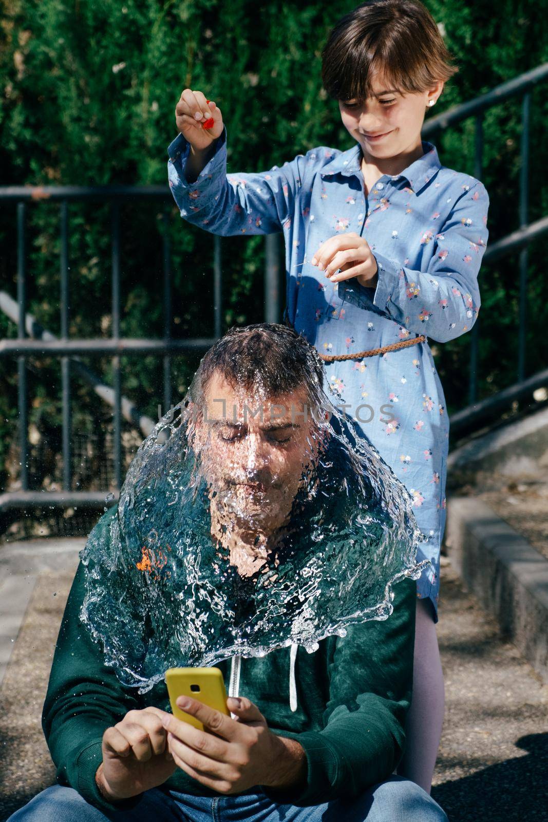 Little girl blowing up a water-filled balloon over her father's head to get him to stop staring at his smartphone.