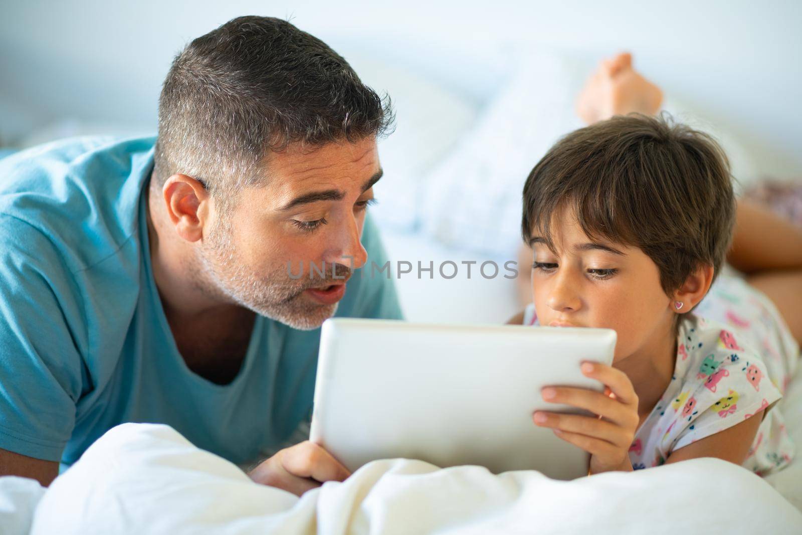 Middle-age father with her eight years daughter using digital tablet in bedroom. Funny dad and lovely little girl are having fun on the bed.