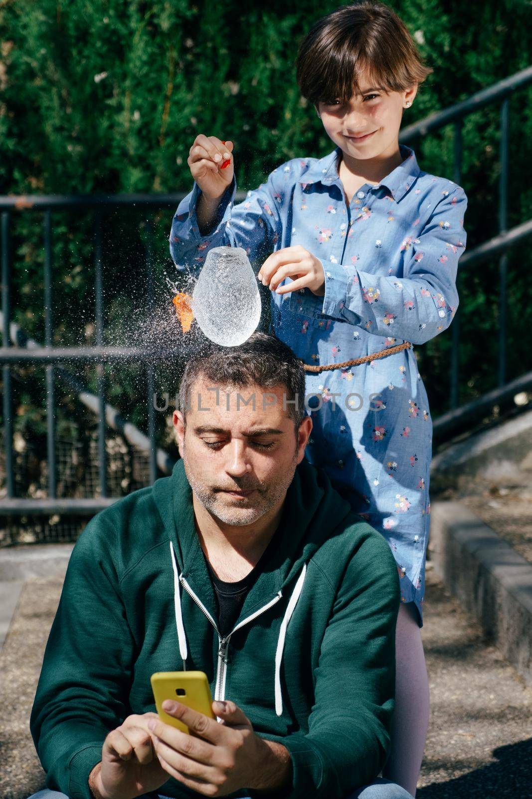 Little girl blowing up a water-filled balloon over her father's head to get him to stop staring at his smartphone.