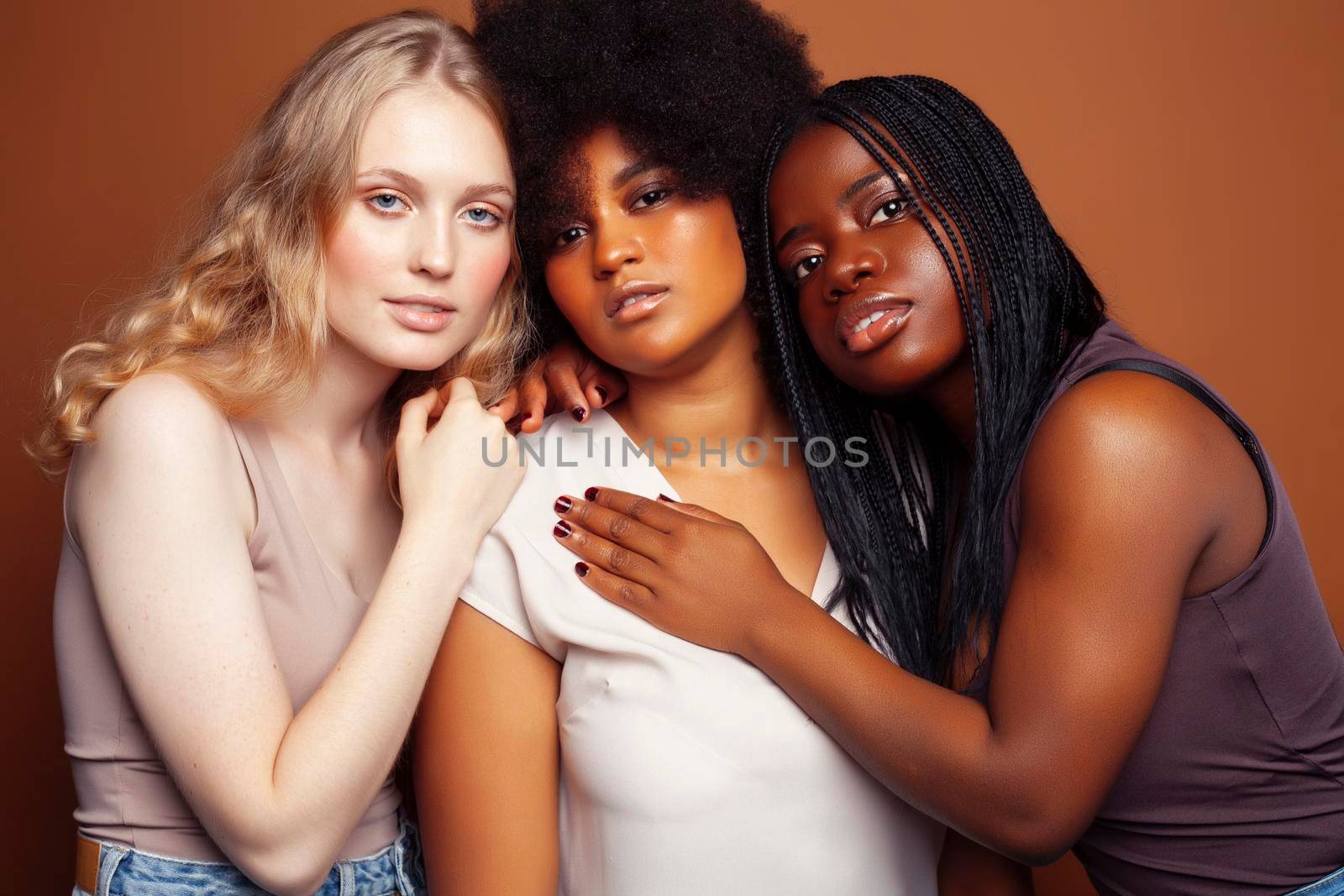 young pretty caucasian, afro, scandinavian woman posing cheerful together on brown background, lifestyle diverse nationality people concept close up
