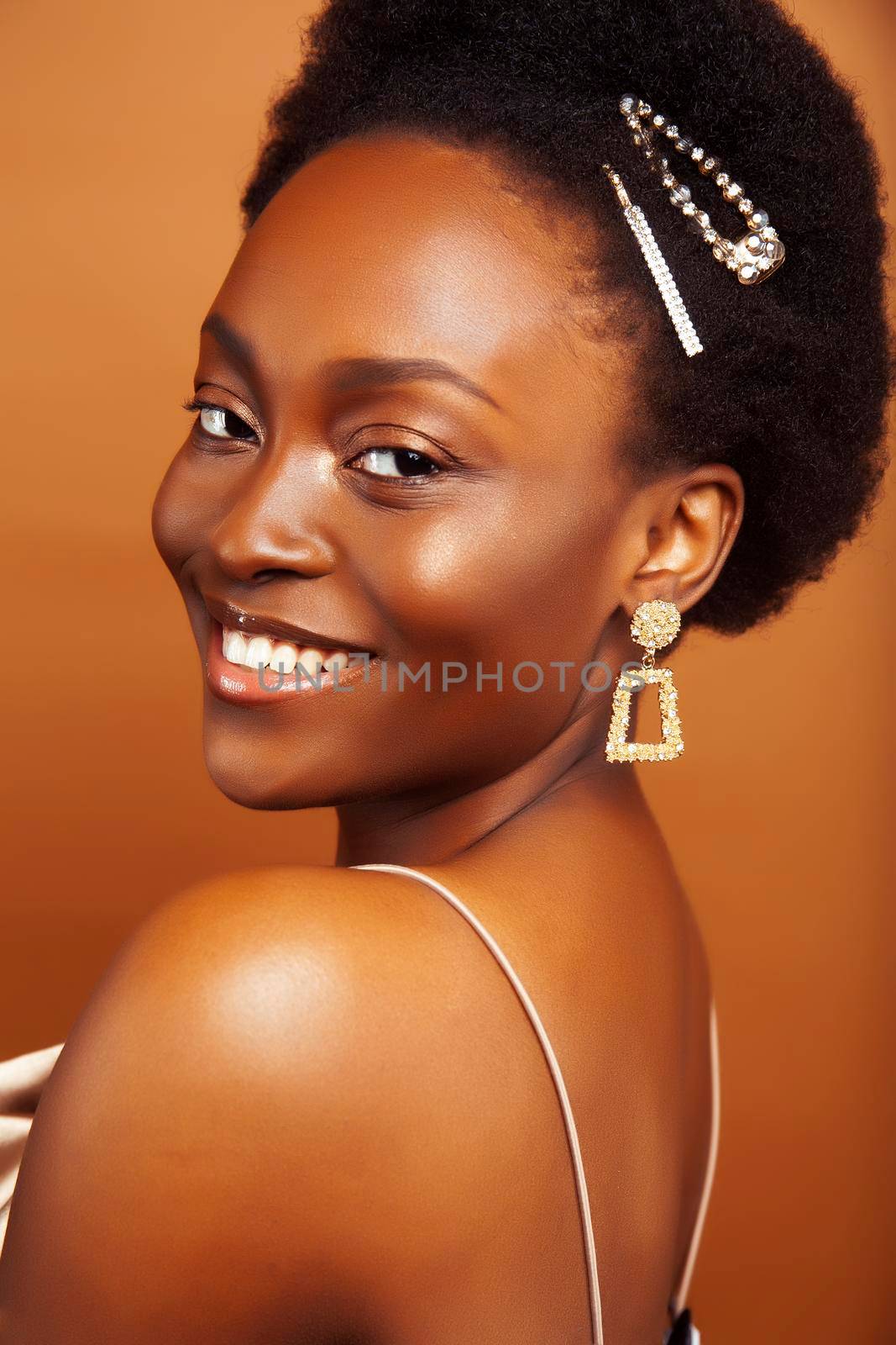 young pretty african model with golden jewelry in fashion style dress smiling happy on brown backround, lifestyle people concept close up
