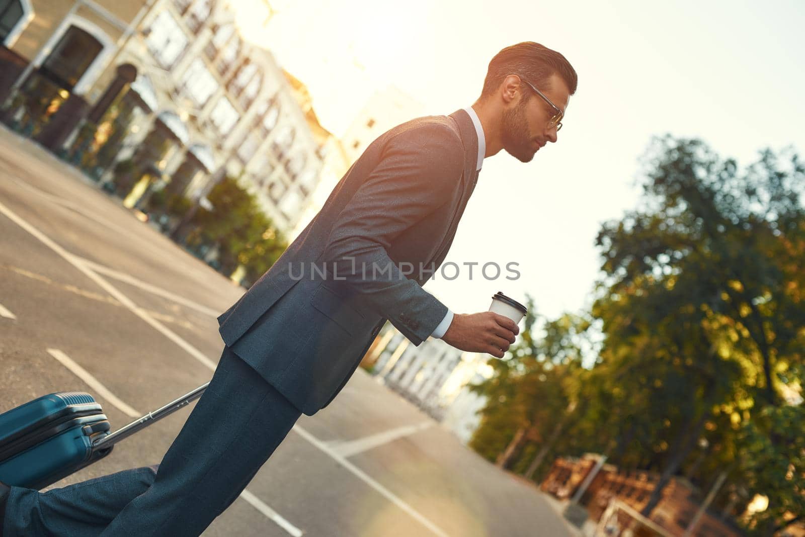 Morning city. Full length of young and handsome bearded businessman in suit pulling his luggage and holding cup of coffee while walking outdoors. Travelling. Business concept