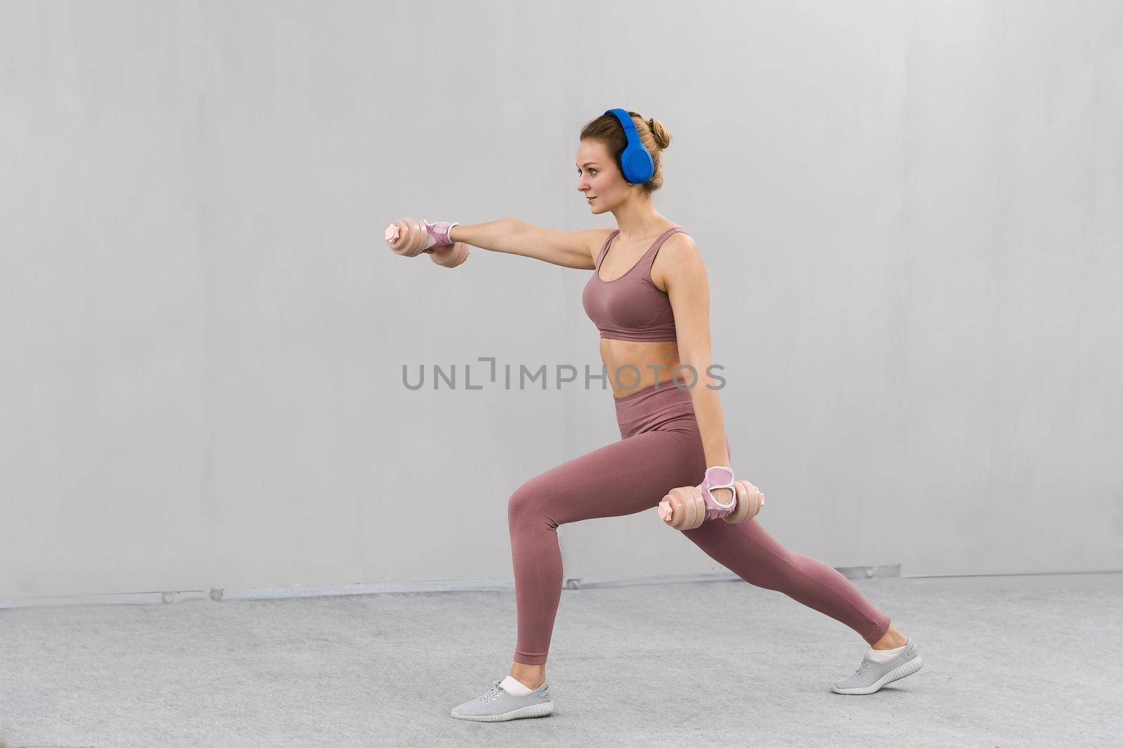 Beautiful fitness girl training legs and lifting weights. Girl in a tight pink outfit working out with dumbbells. Girl doing sports listen music in blue wireless headphones. Sport concept by LipikStockMedia