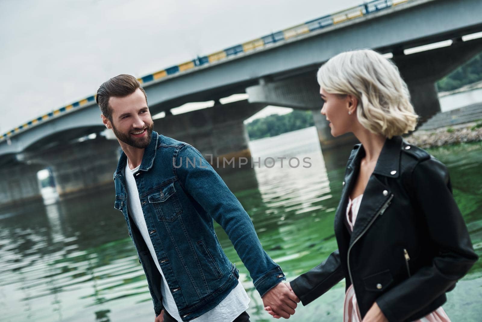 Romantic date outdoors. Young couple walking on the city street near water holding hands looking at each other talking smiling joyful