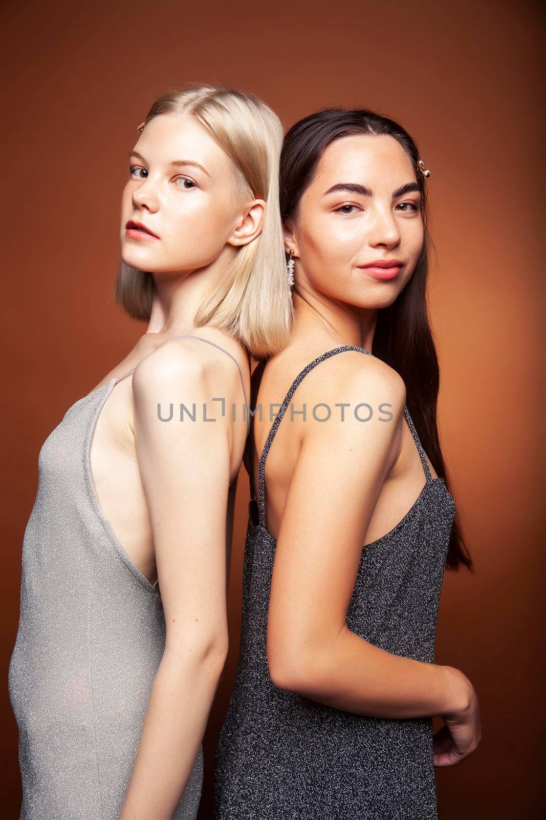 two pretty diverse girls happy posing together: blond and brunette on brown background, lifestyle people concept closeup