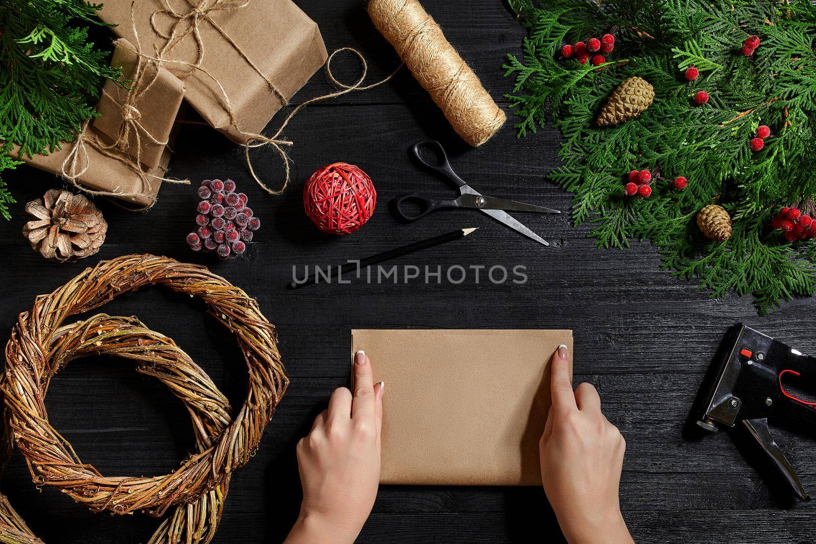 Christmas background. Top view of female hands wrap New Year present. Packed gifts and scrolls, spruce branches and tools on wooden table. Workplace for preparing handmade decorations.