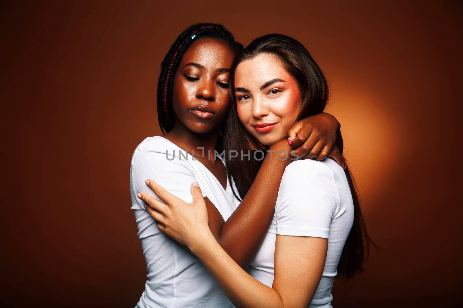 young pretty caucasian, afro woman posing cheerful together on brown background, lifestyle diverse nationality people concept closeup