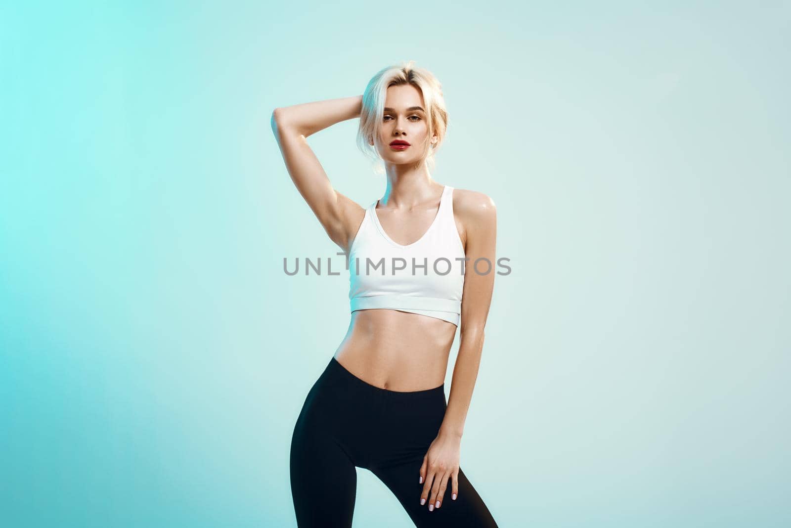 Feeling perfect. Sexy young woman in white top and black leggings touching her blond hair and looking at camera while standing against blue background in studio. Fashion concept. Sensuality. Studio shot