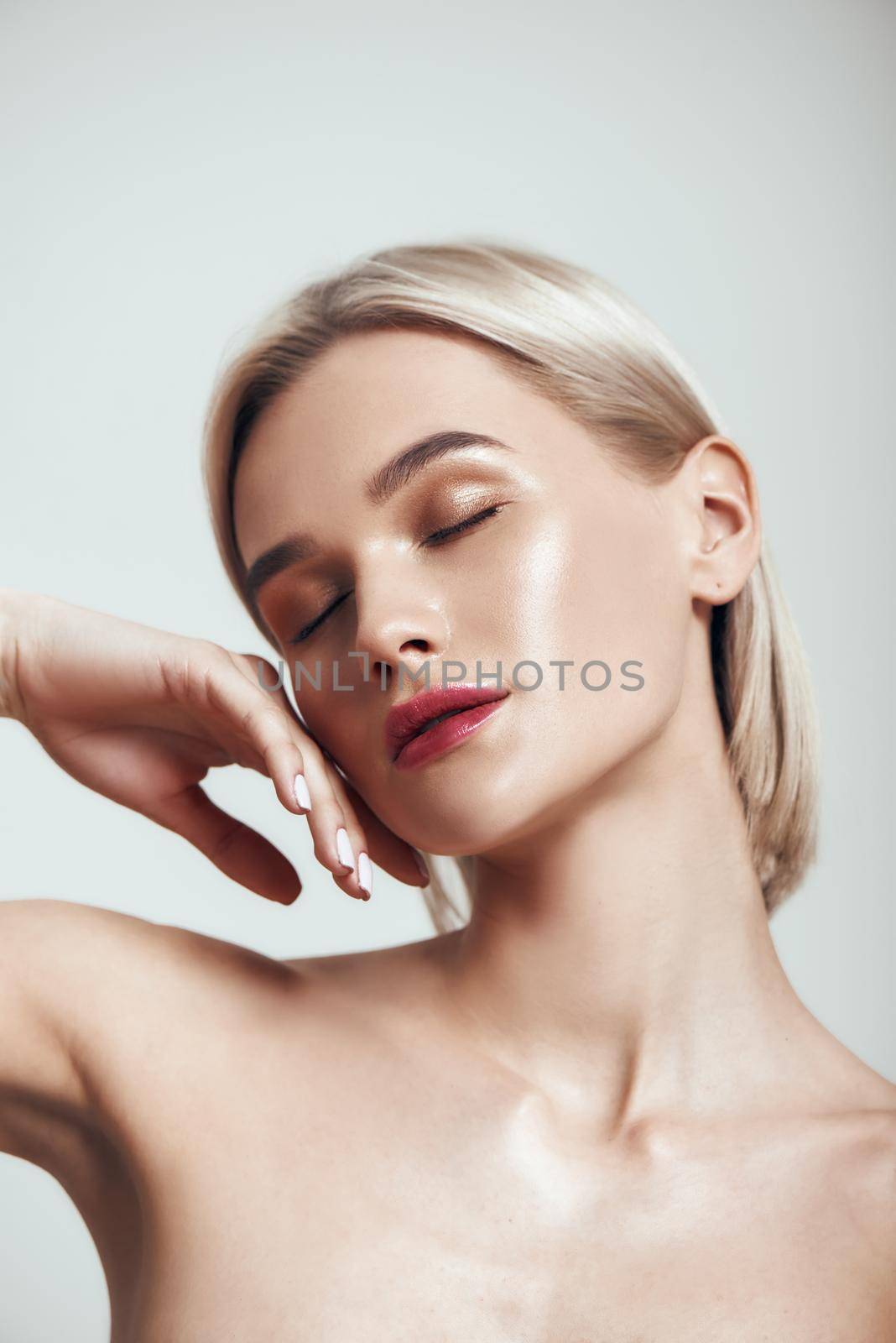 Sensual beauty. Cute young woman with blond hair and perfect shiny skin keeping eyes closed and touching her face while standing against grey background. Women beauty. Skin care. Studio shot