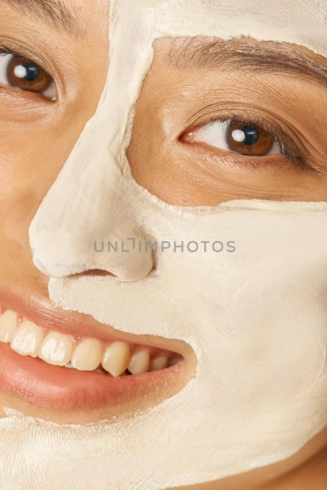 Happy young woman with facial mask applied on half of her face receiving spa treatments, smiling at camera. Beauty, skincare concept