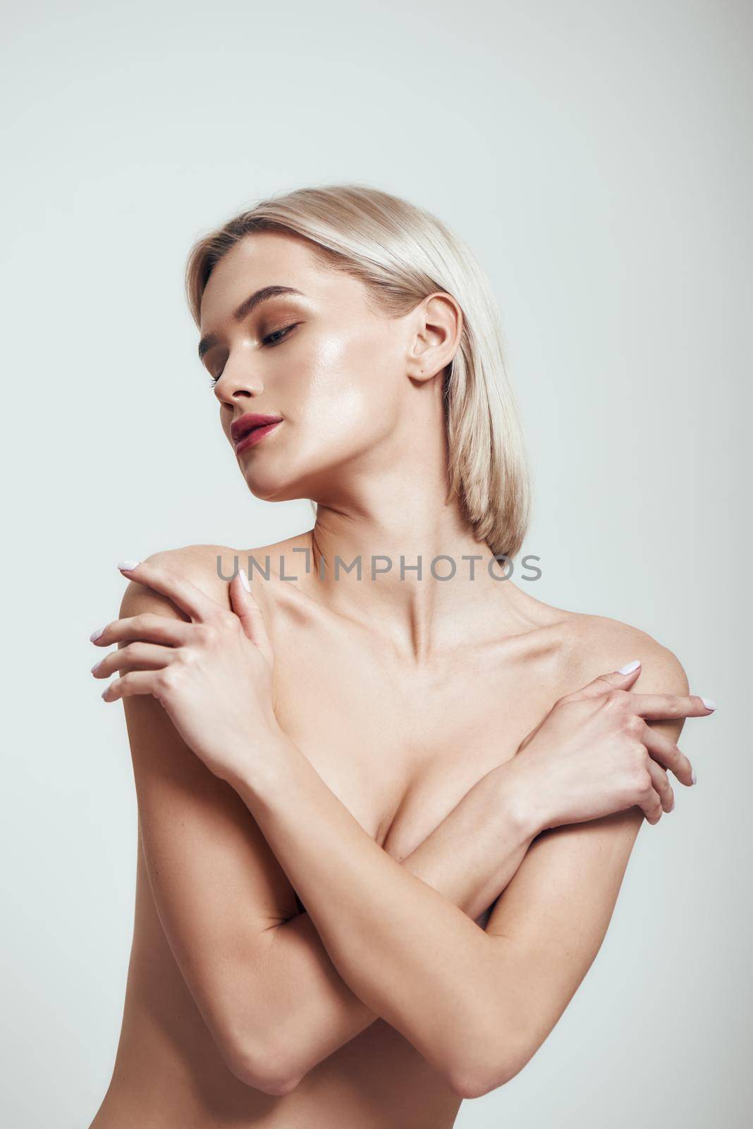 Nude lady. Sexy slim woman with blond hair covering her breast with her hands while standing against grey background. Nude portrait. Sensuality. Beauty concept