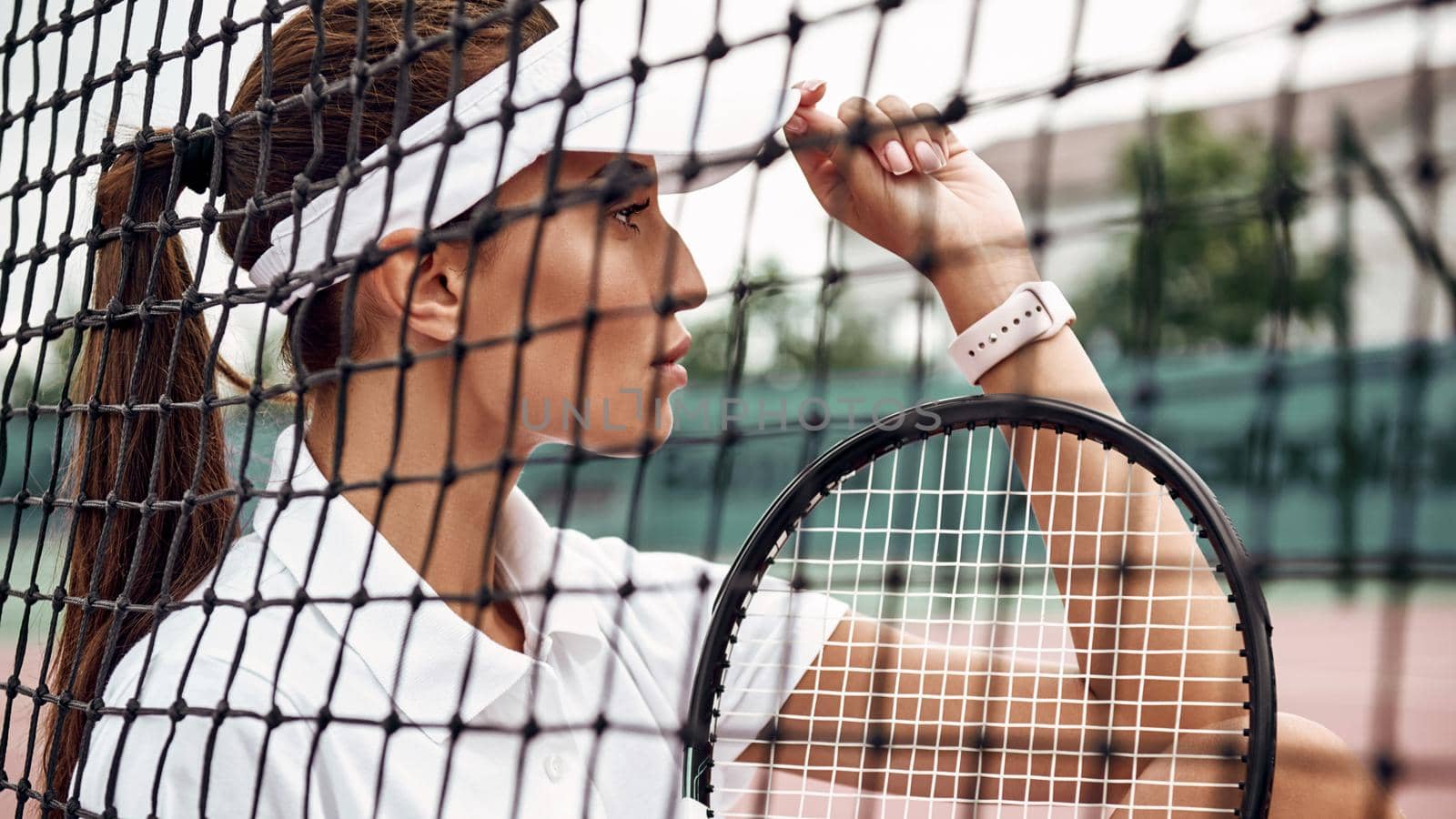 Beautiful sportswoman sitting with a racket on a court, looking away. Healthy lifestyle. A view through tennis net. Horizontal shot