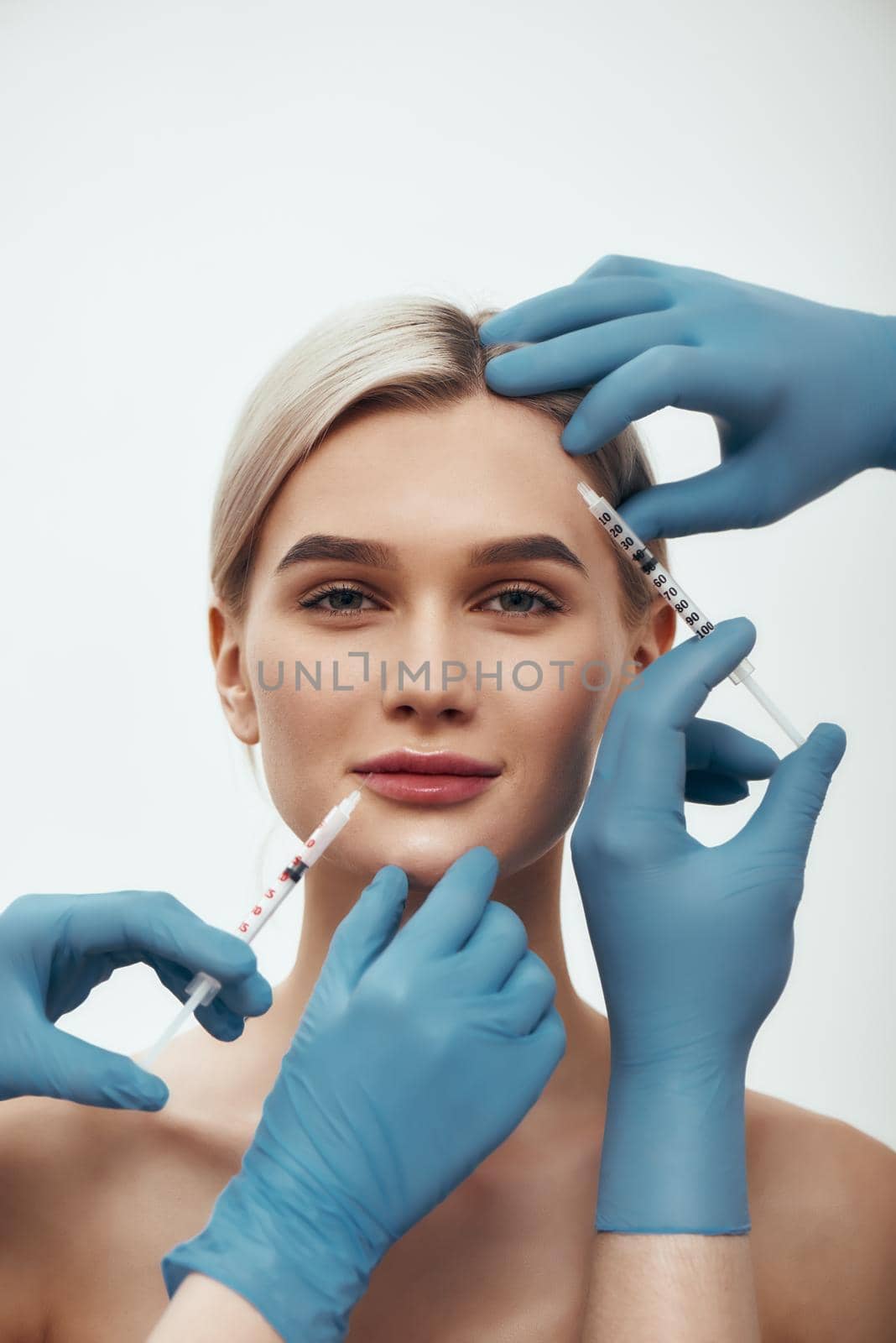 Creating beauty. Portrait of young pretty woman looking at camera and smiling while doctors in blue medical gloves making injections in her face by friendsstock