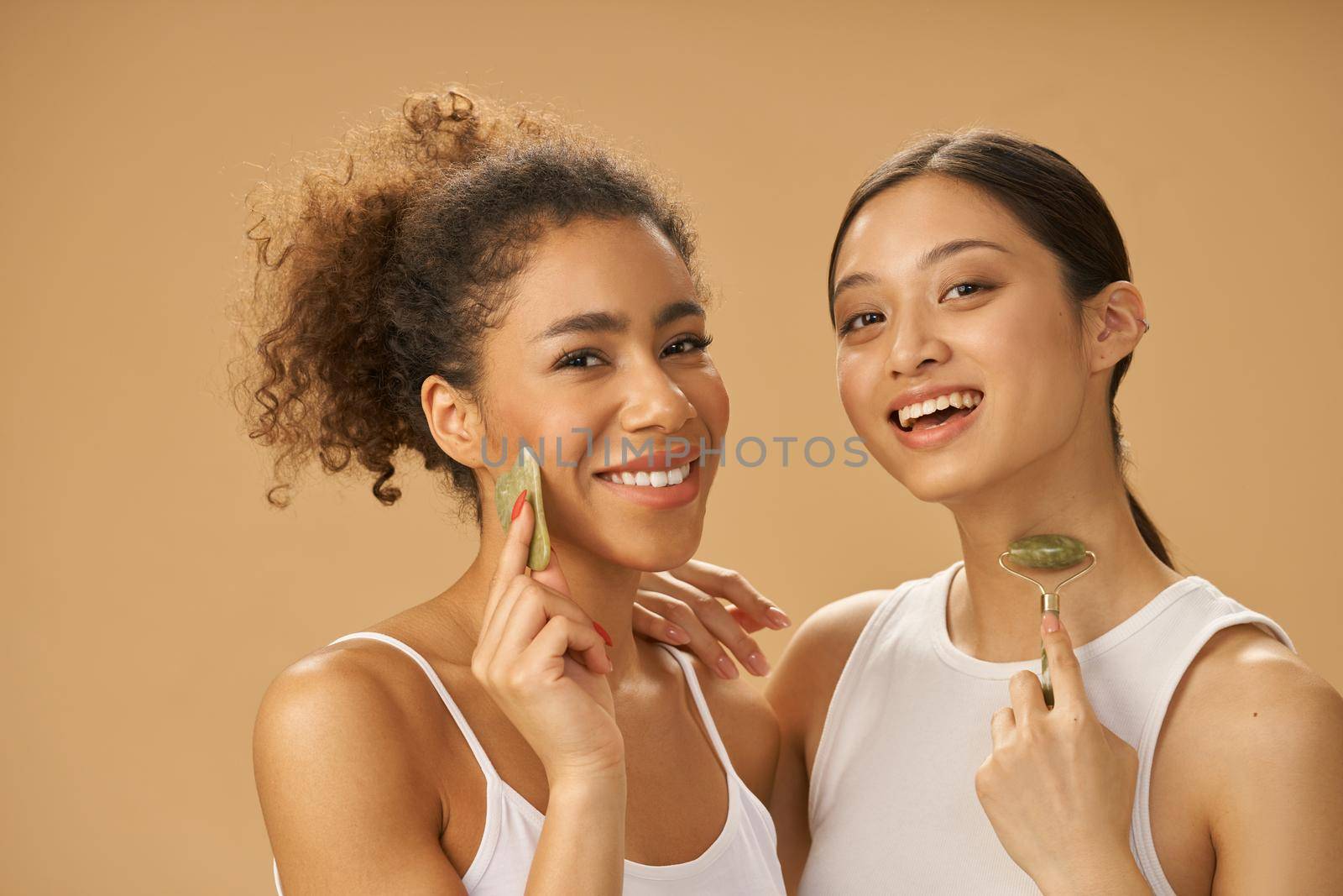 Pretty young women smiling at camera, using jade roller and facial gua sha while posing together isolated over beige background. Skincare concept