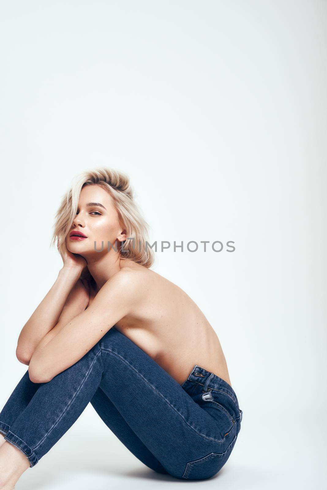 Sensuality. Sexy young woman in jeans covering breasts with hands and looking at camera while sitting on the floor against grey background in studio. Denim clothes. Fashion concept. Sensuality