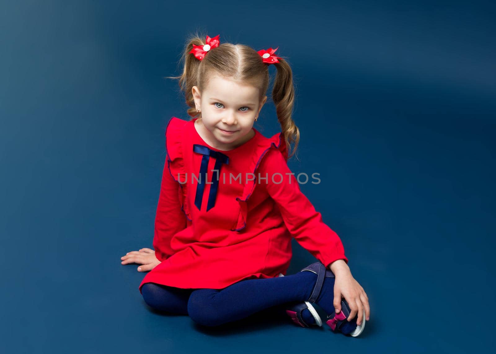 Cute gray eyed girl in red dress sitting on floor. Adorable little girl with cute two ponytails with bows dressed elegant fashionable clothing sitting on floor and looking at something with curiosity