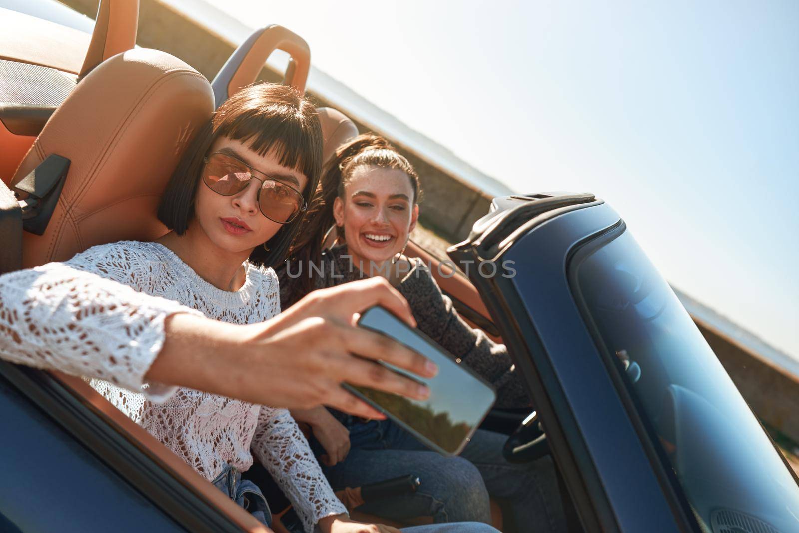 Two happy women in the cabriolet driving, having fun and taking selfie by friendsstock