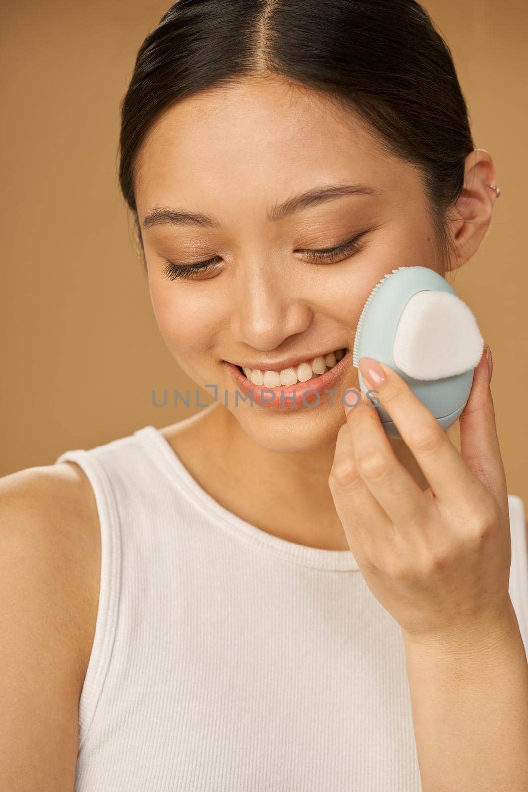 Pretty young woman smiling while using silicone facial cleansing brush, posing isolated over beige background. Washing accessory. Skincare concept