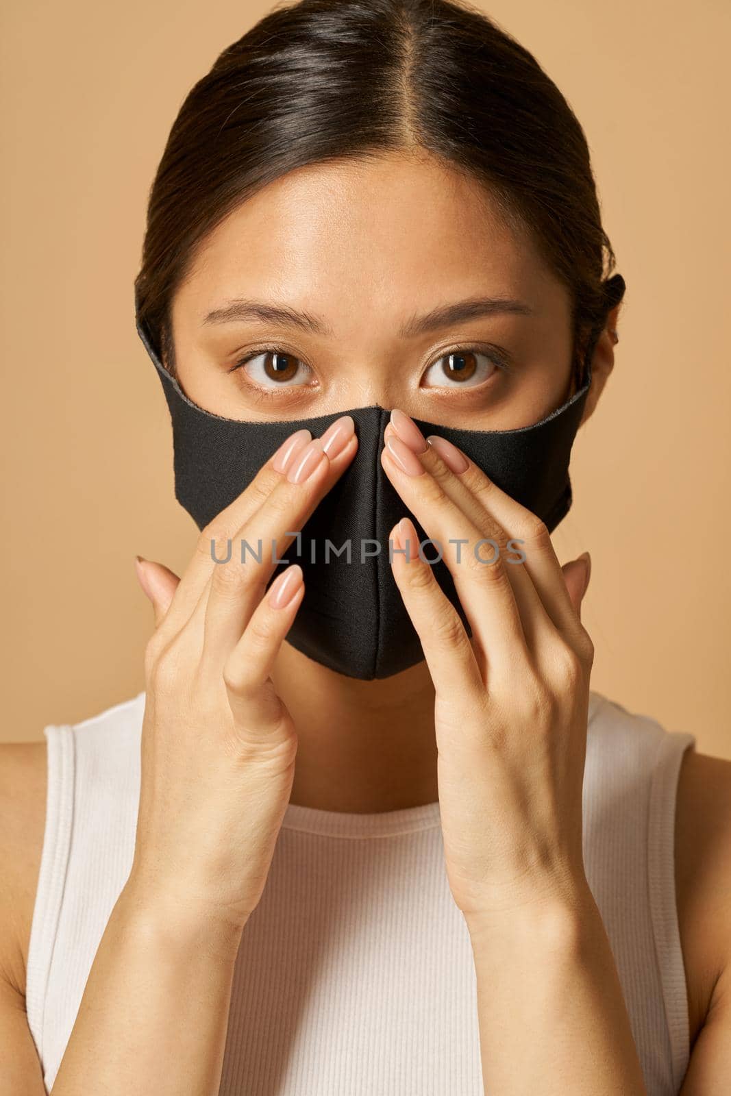 Studio portrait of young woman putting on black facial mask, looking at camera while posing isolated over beige background by friendsstock