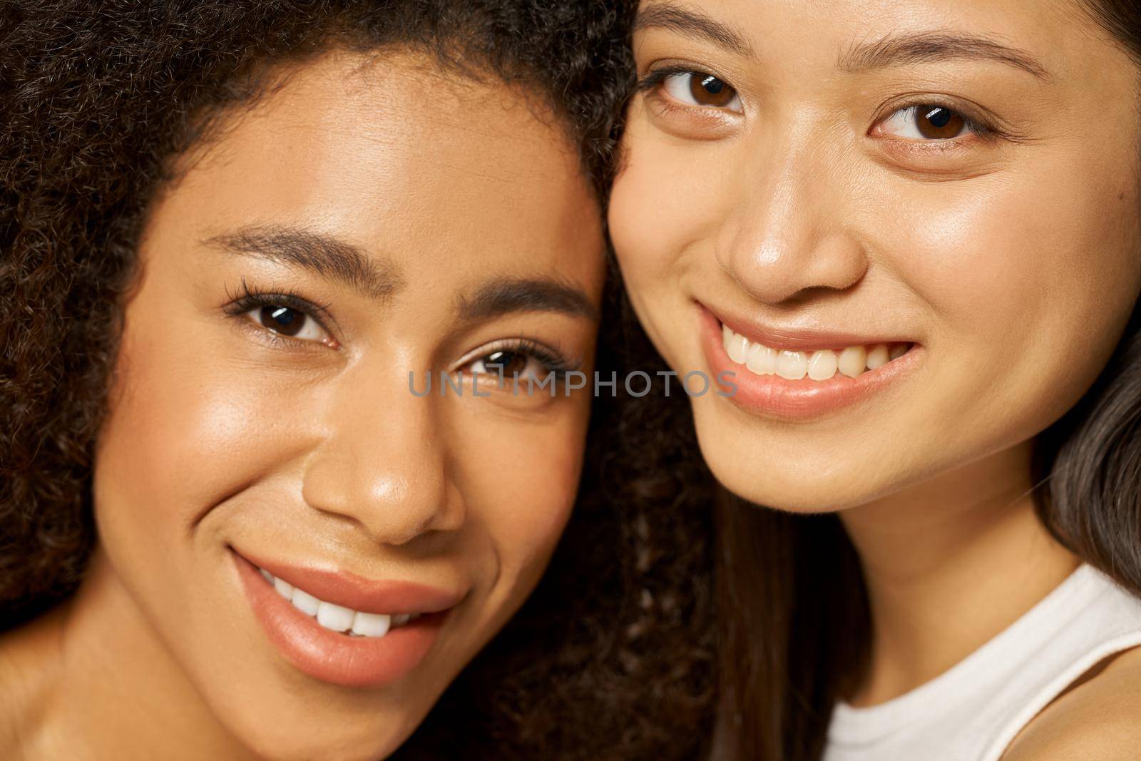 Face closeup of two beautiful happy mixed race young women with perfect glowing skin smiling at camera while posing together. Skincare, diversity concept. Selective focus. Horizontal shot