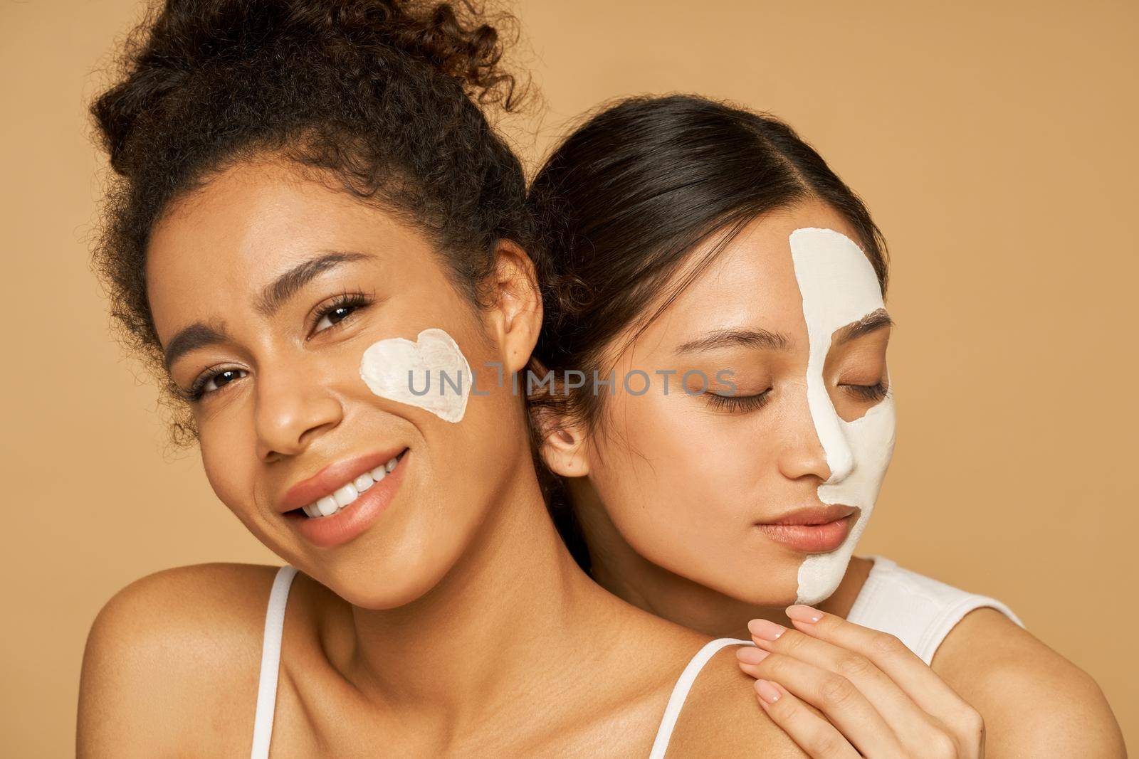 Two attractive young women with facial masks on posing isolated over beige background. Skincare, beauty concept. Horizontal shot