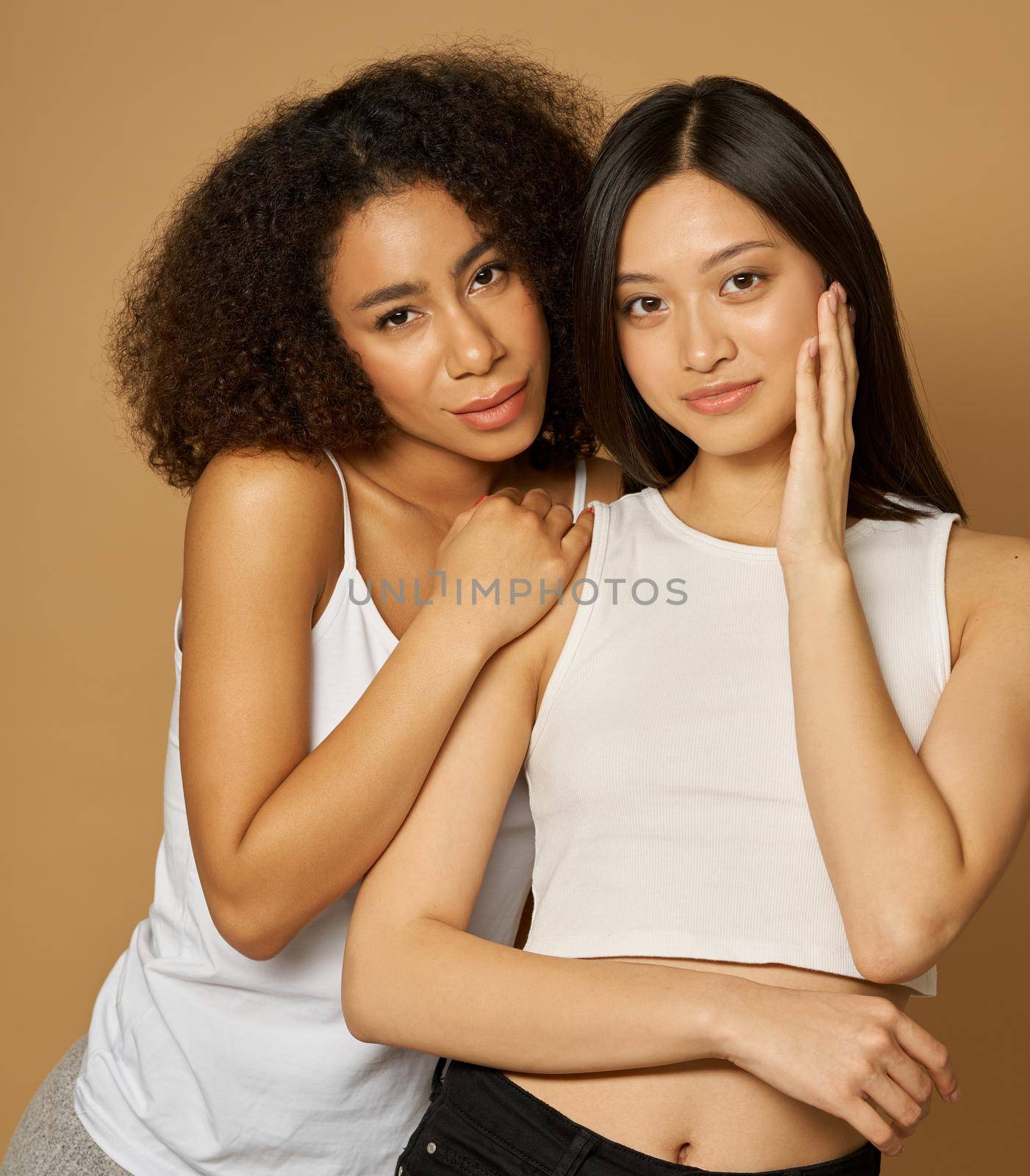 Portrait of two attractive mixed race young women looking at camera while posing together, standing isolated over light brown background. Skincare, diversity concept