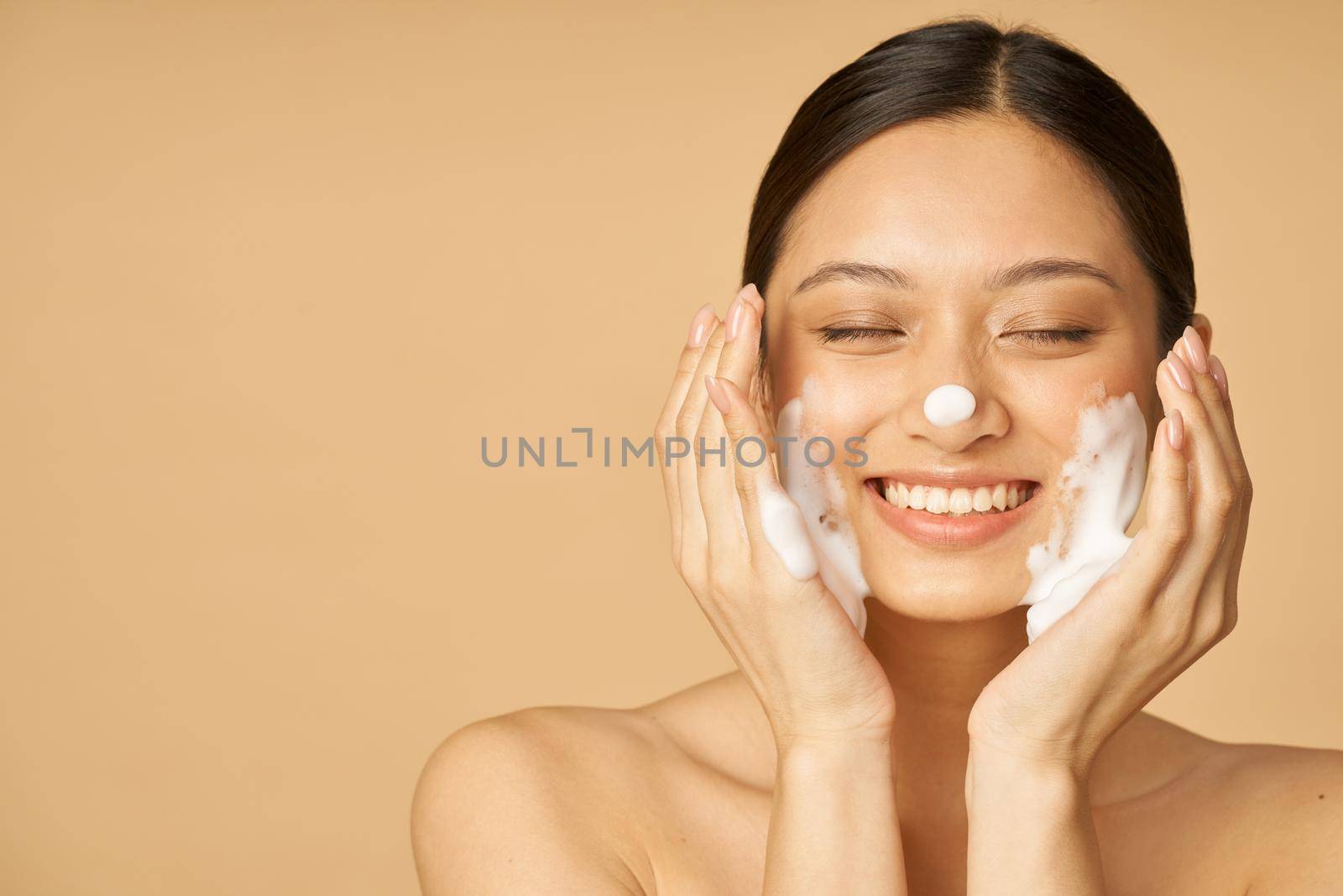 Studio portrait of pleased young woman smiling with eyes closed while applying gentle foam facial cleanser isolated over beige background. Beauty products and skin care concept. Front view