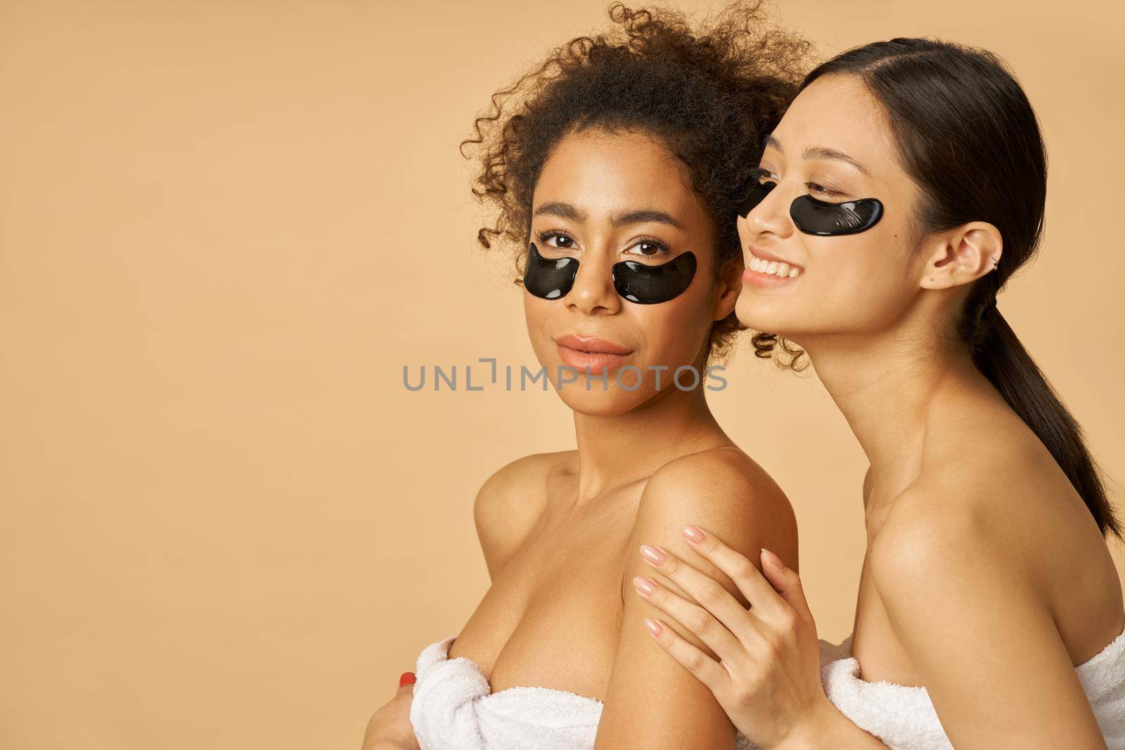 Beauty portrait of two cheerful young women posing with applied black hydro gel under eye patches isolated over beige background. Skincare routine concept
