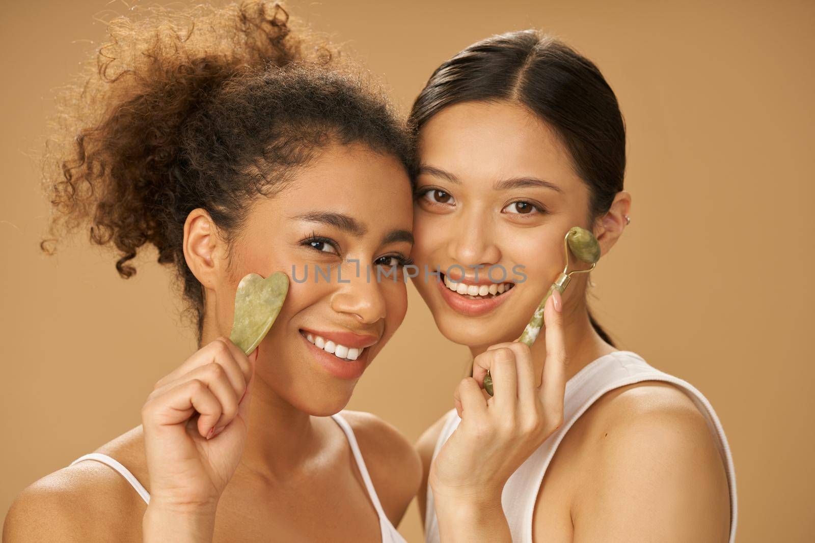 Cute young women smiling at camera, using jade roller and facial gua sha while posing together isolated over beige background. Skincare concept