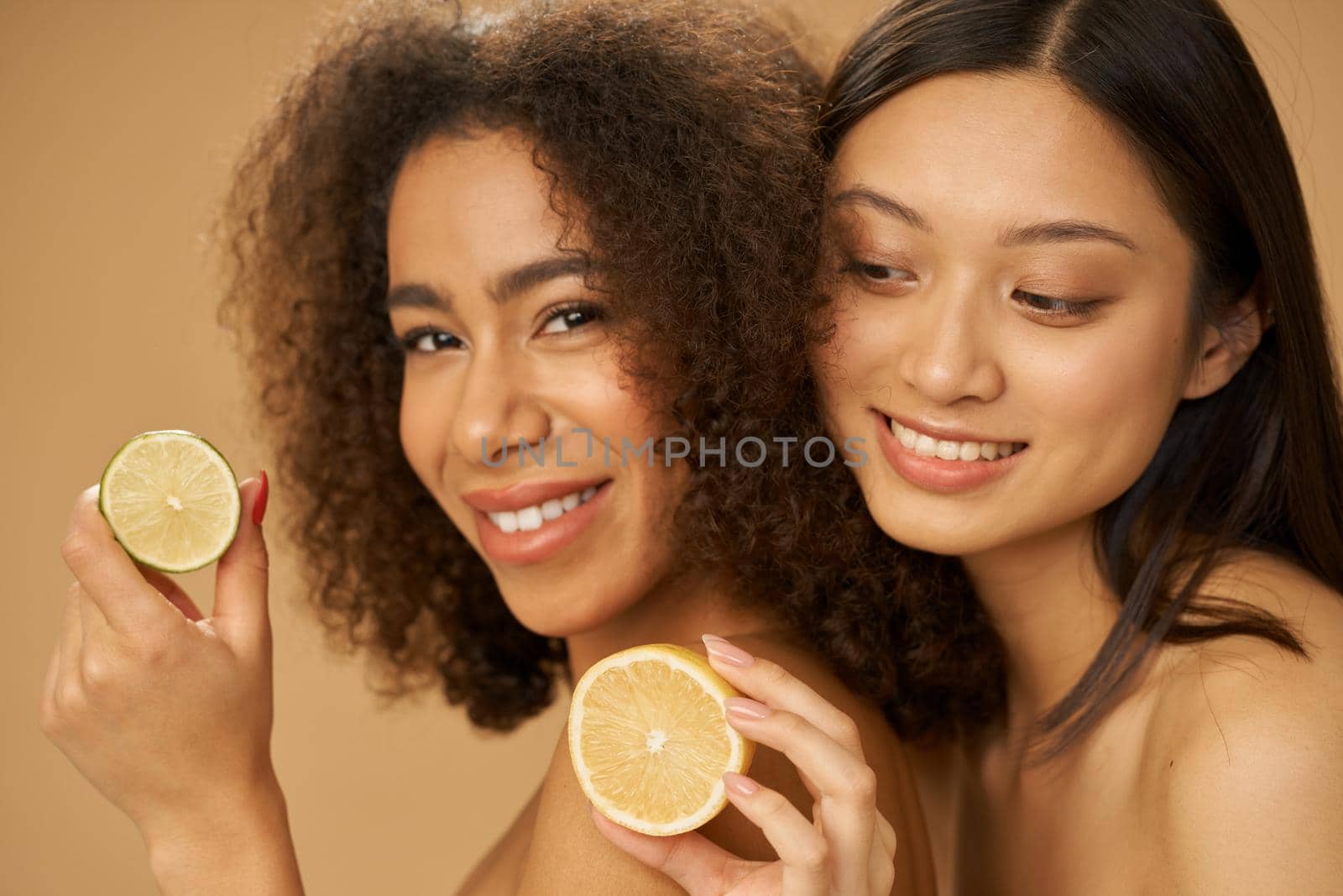 Two lovely mixed race young women looking excited, holding lemon and lime cut in half while posing together isolated over beige background. Health and beauty concept