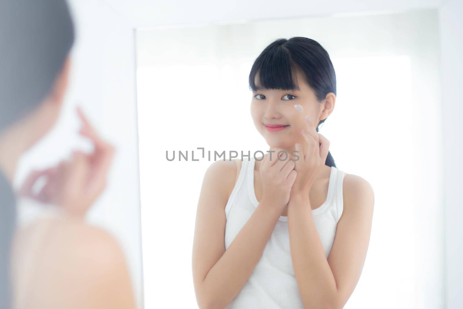Beautiful young asian woman looking in mirror and apply moisturizer with cosmetic cream or lotion in bathroom at home, skincare for hygiene, treatment and bodycare, skin and anti-aging of facial.