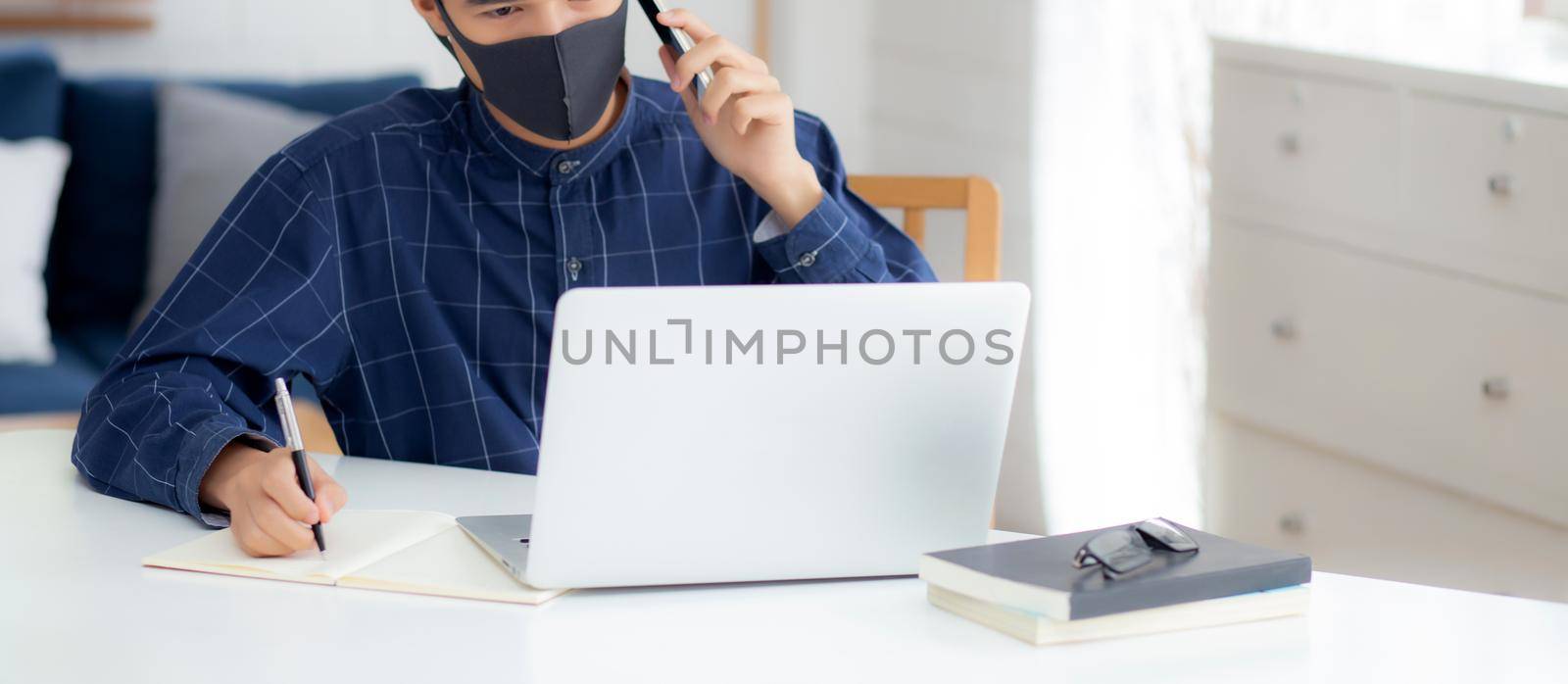 Young asian business man in face mask talking smartphone and work on laptop computer for protect covid-19, businessman quarantine work from home and writing notebook, social distancing and stay home. by nnudoo