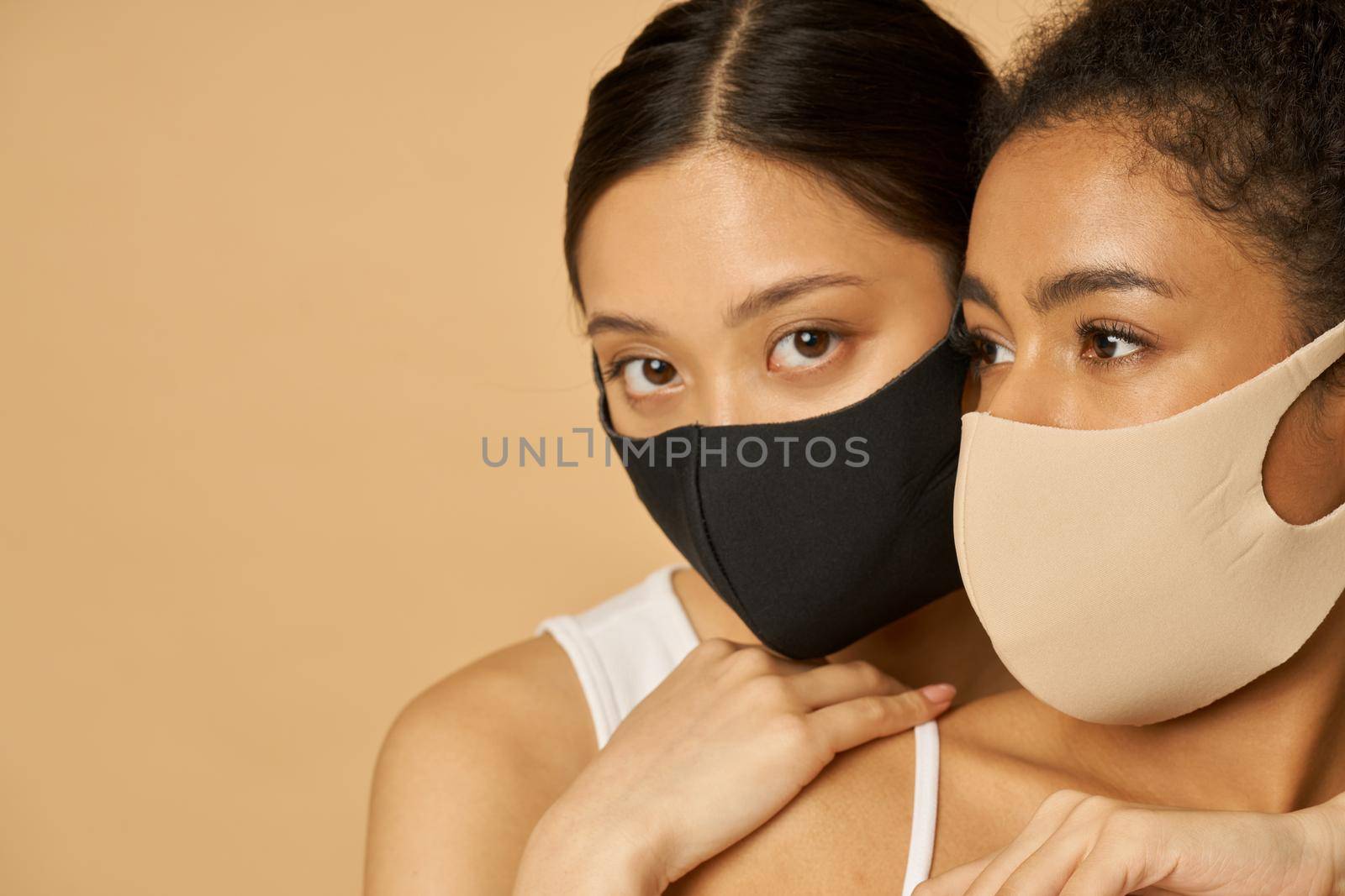 Two adorable young women, female friends wearing protective facial masks while posing together isolated over beige background. Safety, pandemic concept
