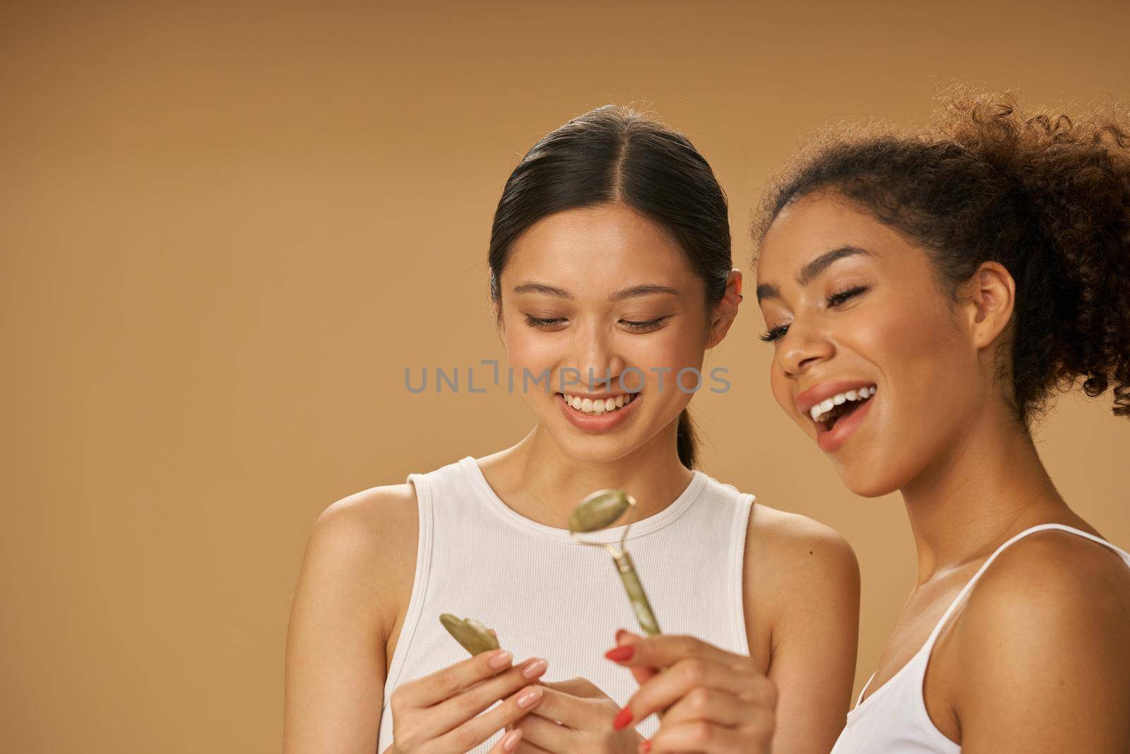 Smiling young women holding, looking at jade roller and facial gua sha while posing together isolated over beige background by friendsstock