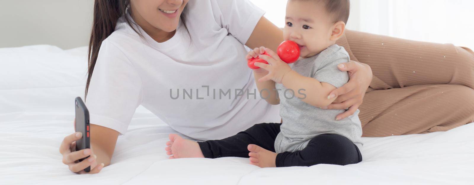 Young asian mother and little baby girl or newborn selfie with smart phone on bed in bedroom, happiness mom and daughter using phone video call at home, two people, family and communication concept. by nnudoo