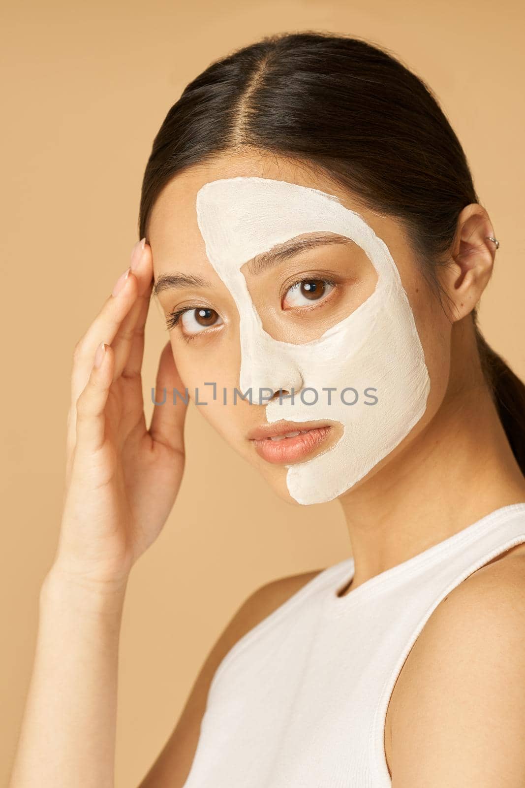 Studio shot of young woman with facial mask applied on half of her face looking at camera, receiving spa treatments isolated over beige background by friendsstock