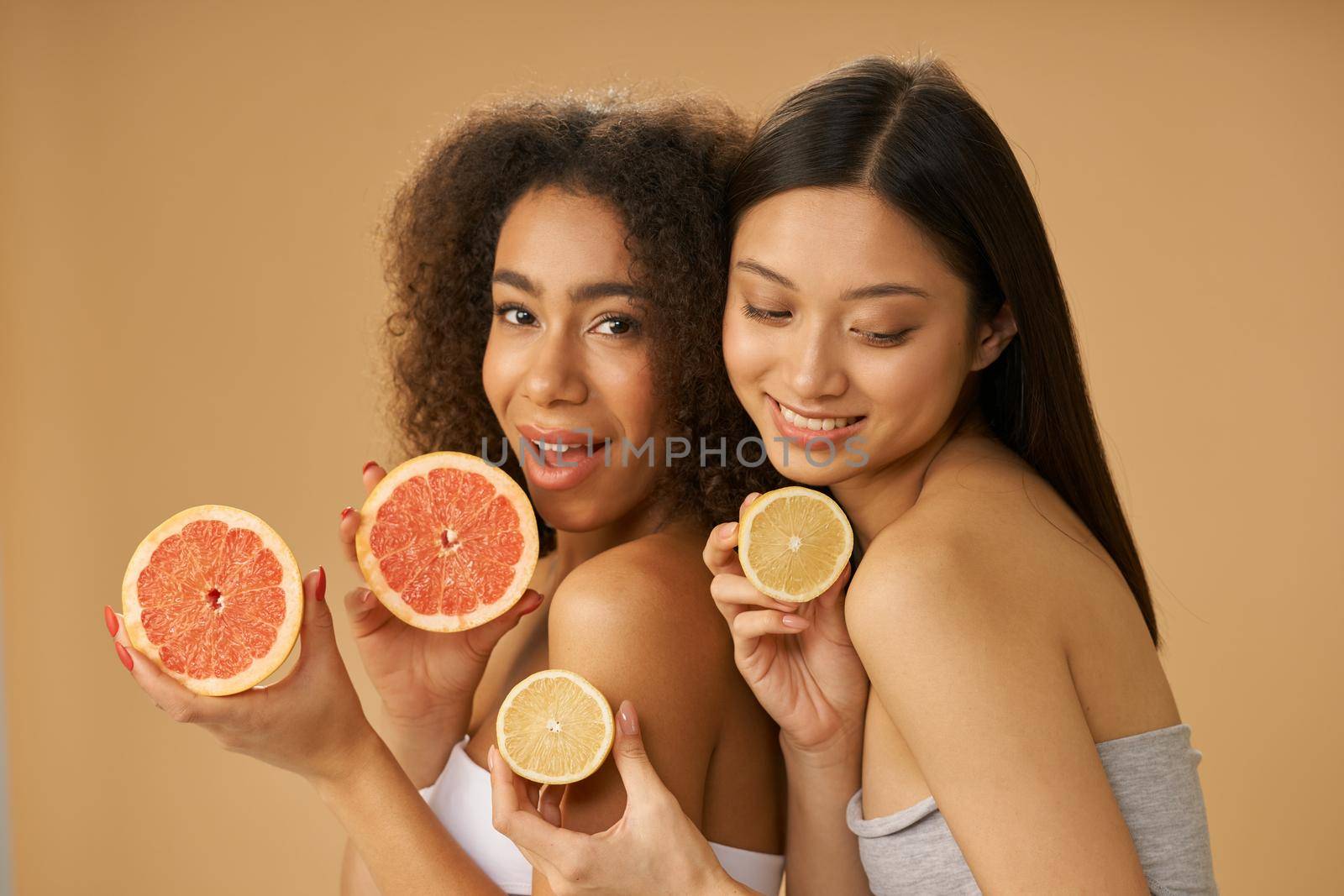 Portrait of two lovely mixed race young women holding grapefruit and lemon cut in half while posing isolated over beige background. Health and beauty concept