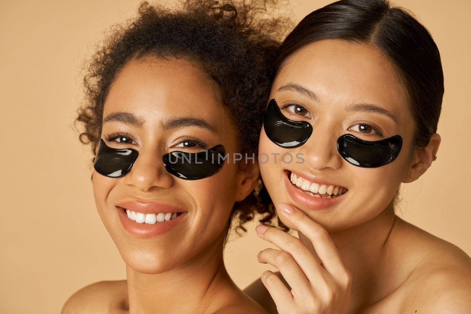 Beauty portrait of two excited young women posing with applied black under eye patches isolated over beige background by friendsstock