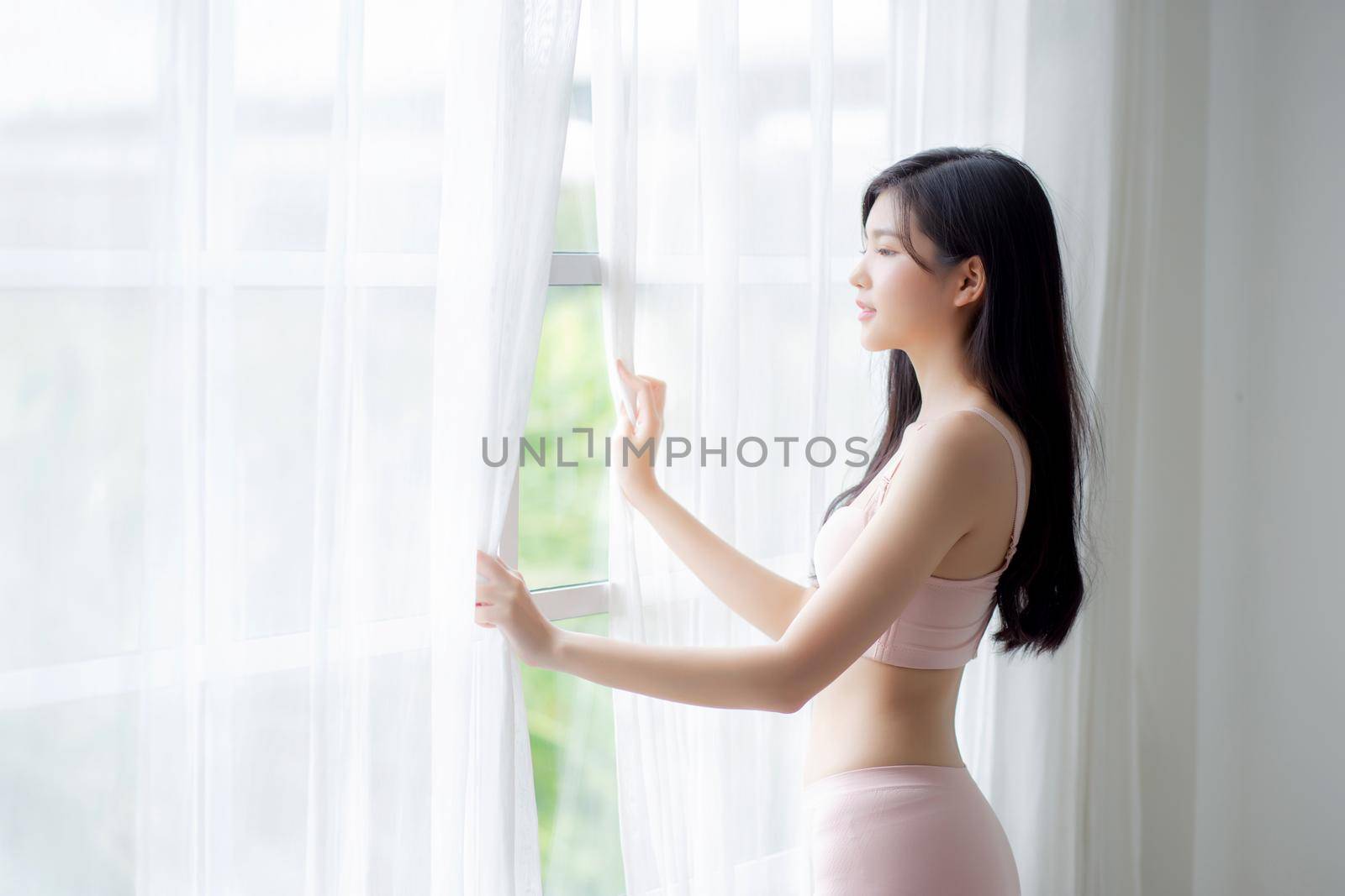 Beautiful young asian woman wake up and walking to window with open curtain in the morning with cozy, sexy girl in underwear smiling relax and resting in the vacation with happy, lifestyle concept. by nnudoo