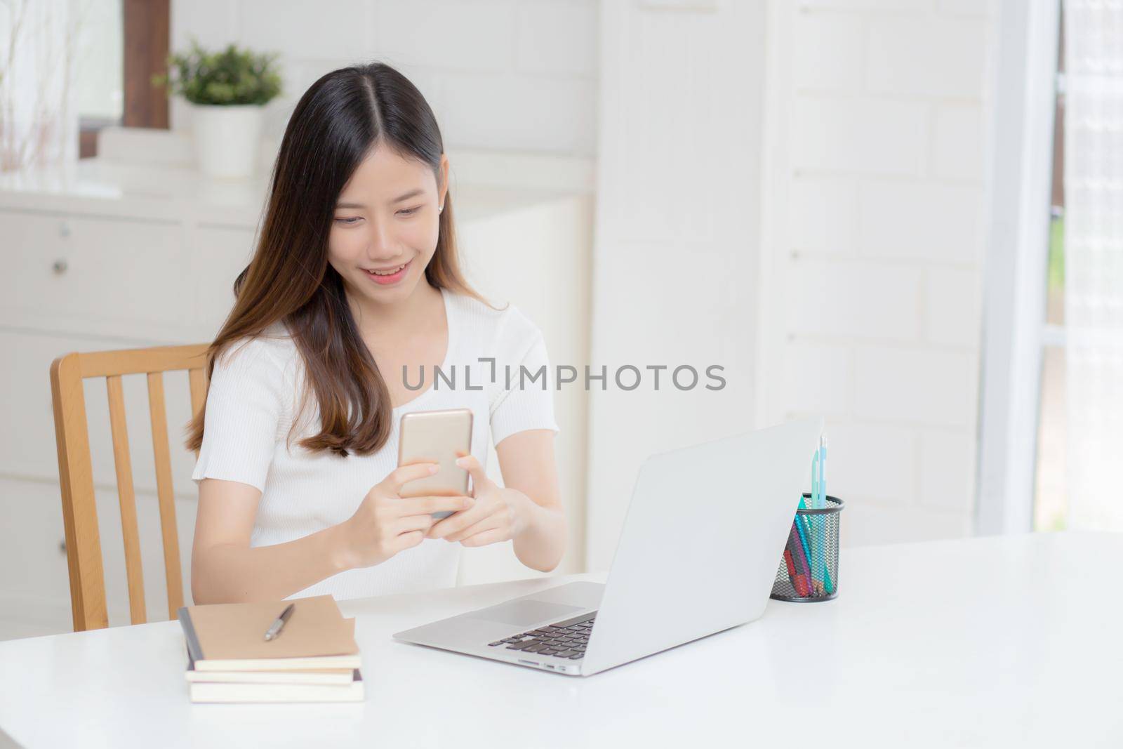 Young asian woman working laptop computer and reading smartphone on internet online on desk at home, freelance girl smiling using phone with social media, business and communication concept. by nnudoo