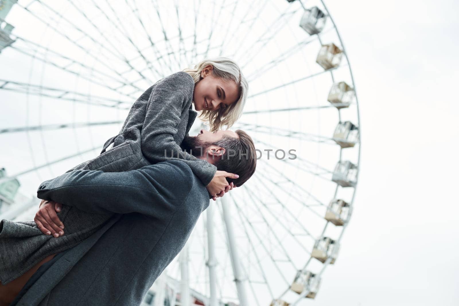 Romantic date outdoors. Young man holding woman up near the ferris wheel looking at each other smiling cheerful by friendsstock