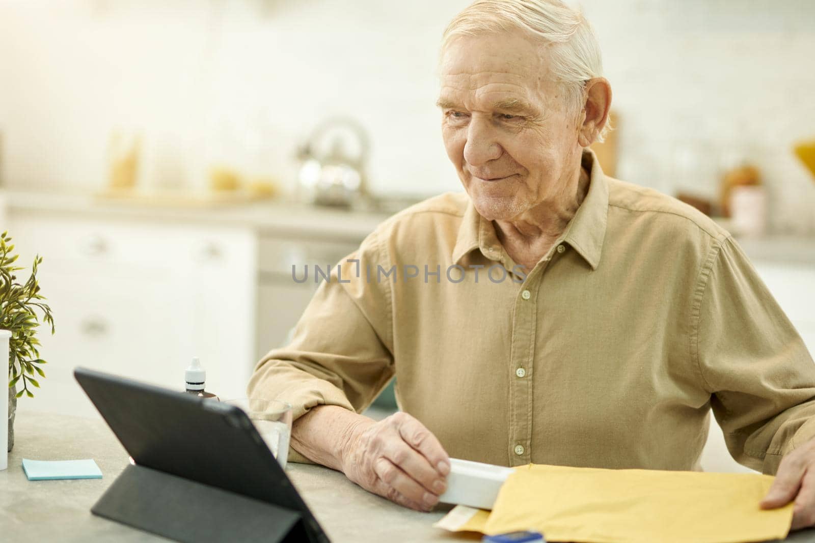 Waist-up photo of an optimistic senior man sitting at the table and looking at the tablet screen