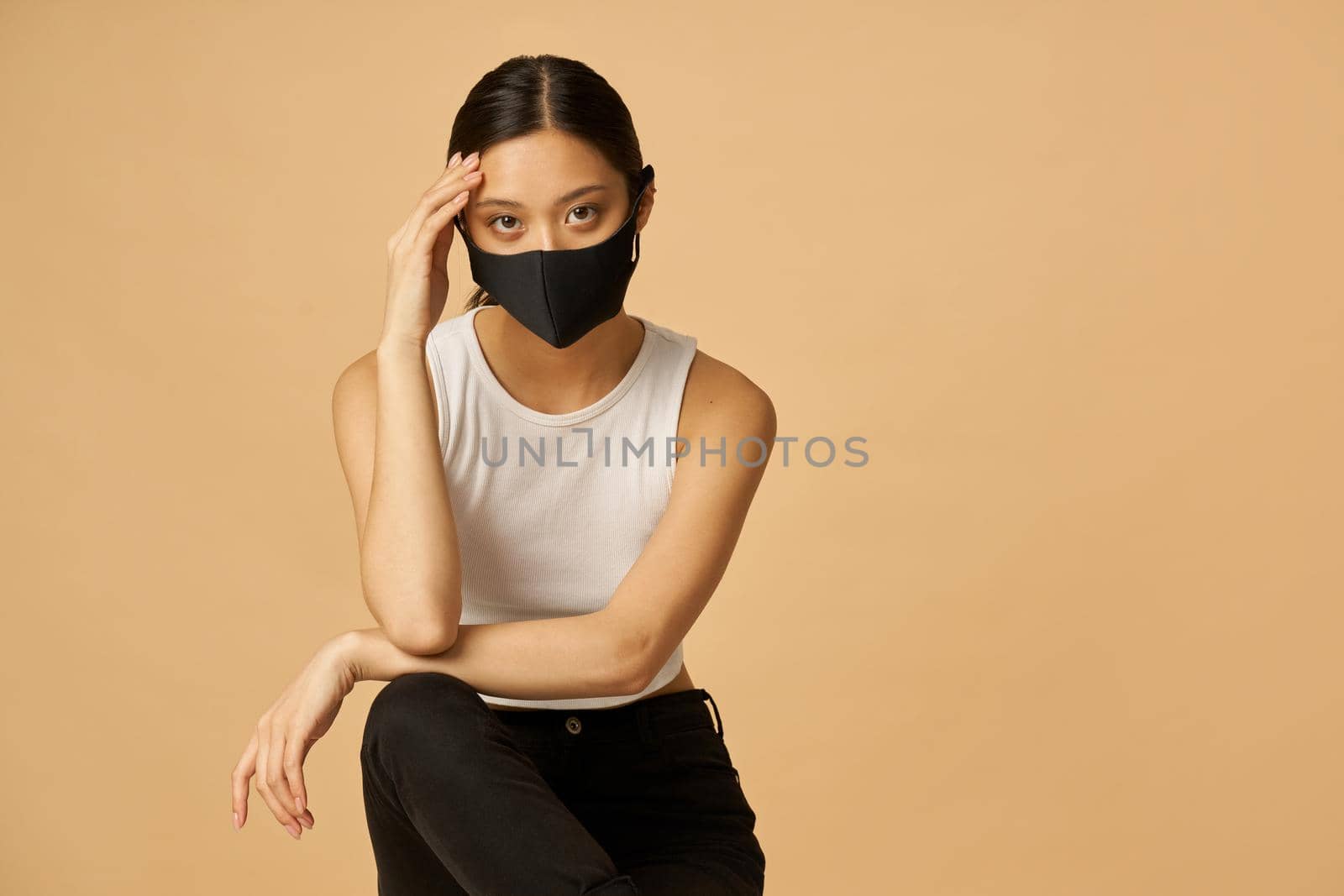 Confident young mixed race woman wearing black facial mask, staying safe, looking at camera while posing isolated over beige background. Safety, pandemic concept