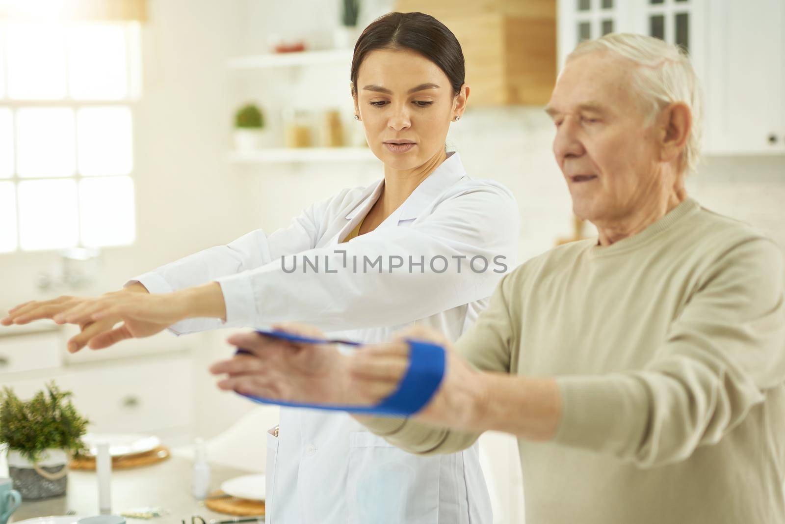 Waist up of medical professional standing near elderly man and showing exercises with fitness rubber band