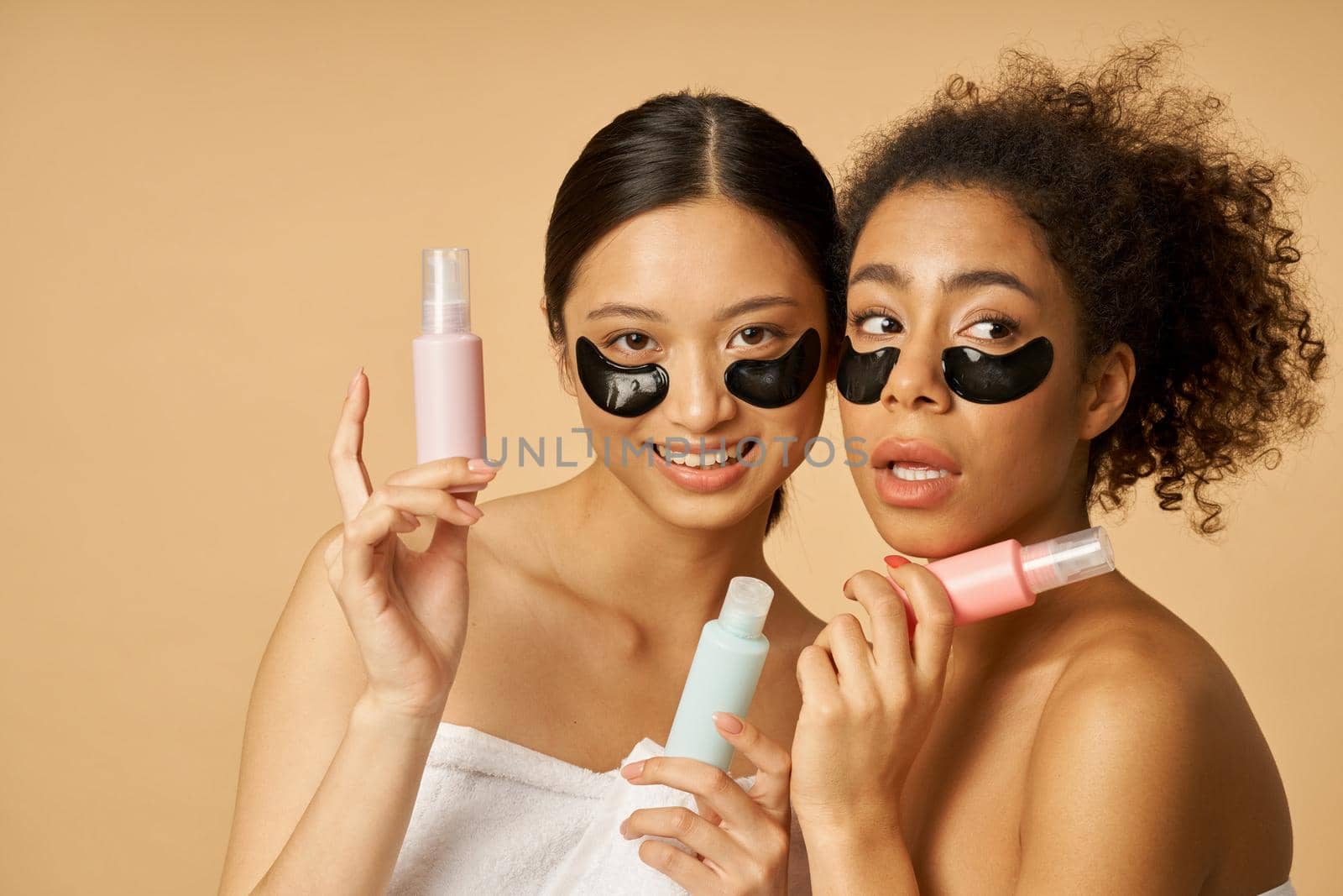 Portrait of two funny young women holding bottles with beauty product, posing with applied black hydro gel under eye patches isolated over beige background by friendsstock